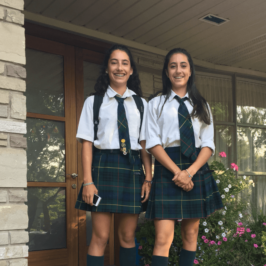 Two teenage girls wearing matching school uniforms—white shirts, plaid skirts, green ties, and knee-high socks—stand and smile outside a building with a wooden door, stone wall, and garden flowers.