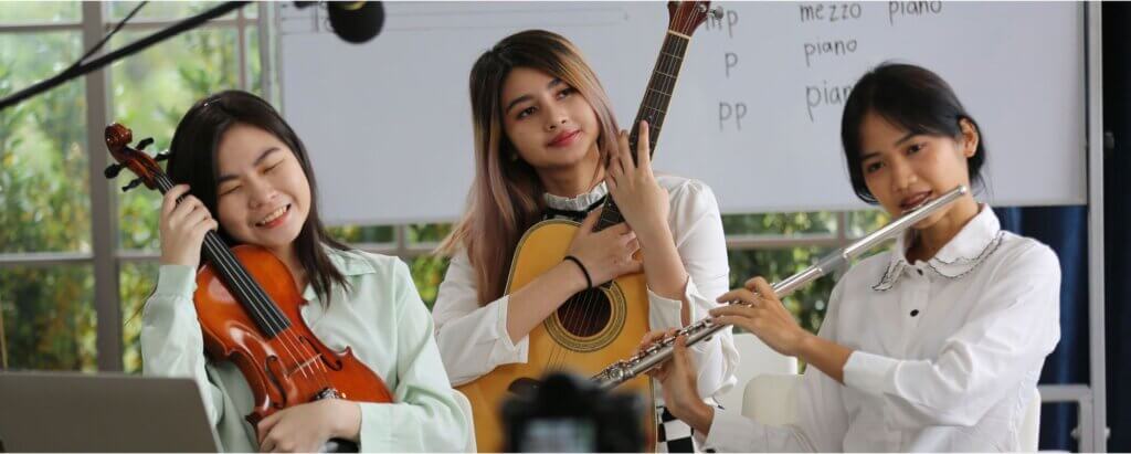 Three young women play musical instruments—violin, guitar, and flute—indoors in front of a whiteboard with music terms written on it. They appear to be performing or practicing together.