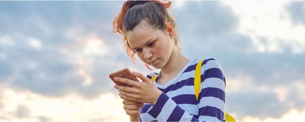 A young person with a ponytail and a yellow backpack looks intently at a smartphone while wearing a blue and white striped shirt, standing outdoors against a cloudy sky.