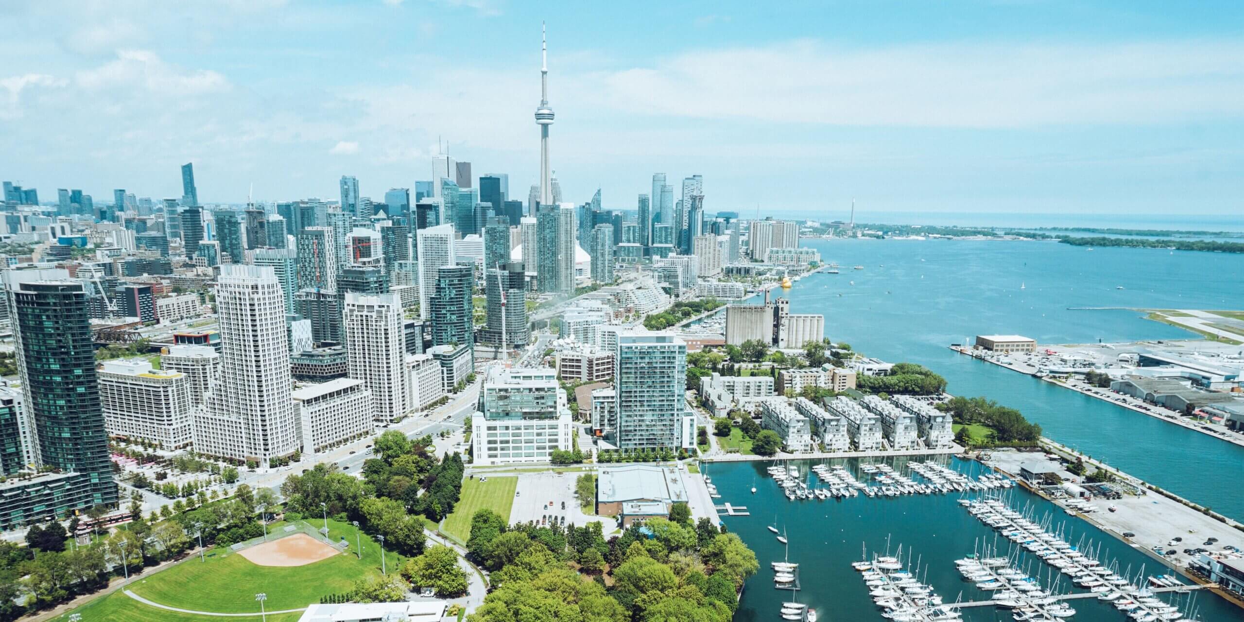 Aerial view of downtown Toronto featuring the CN Tower, high-rise buildings, a marina with boats, green parks, and Lake Ontario under a blue sky.