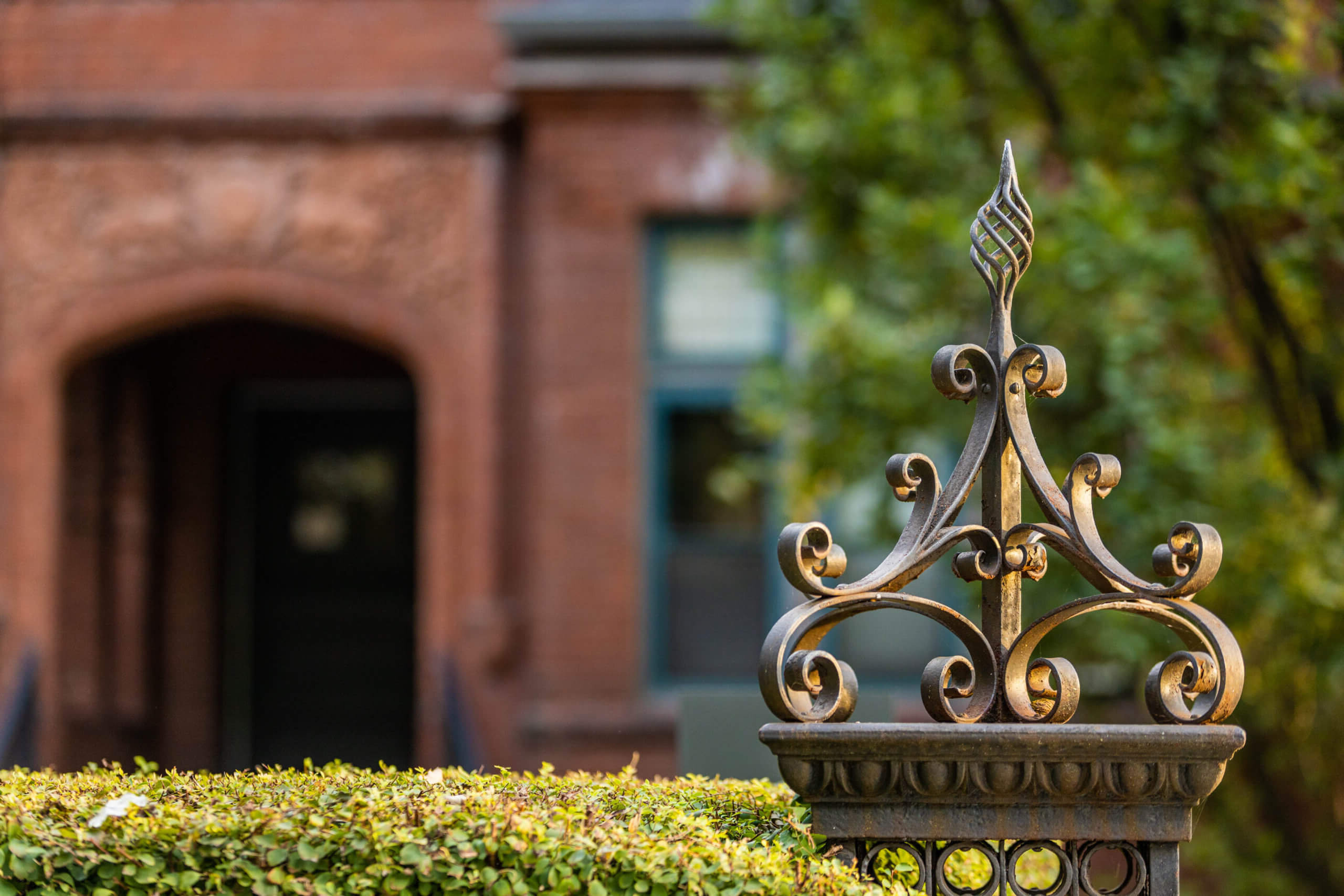 Decorative wrought iron fence post with ornate scrollwork in focus, with a blurred background showing green foliage and the entrance of a red-brick building.
