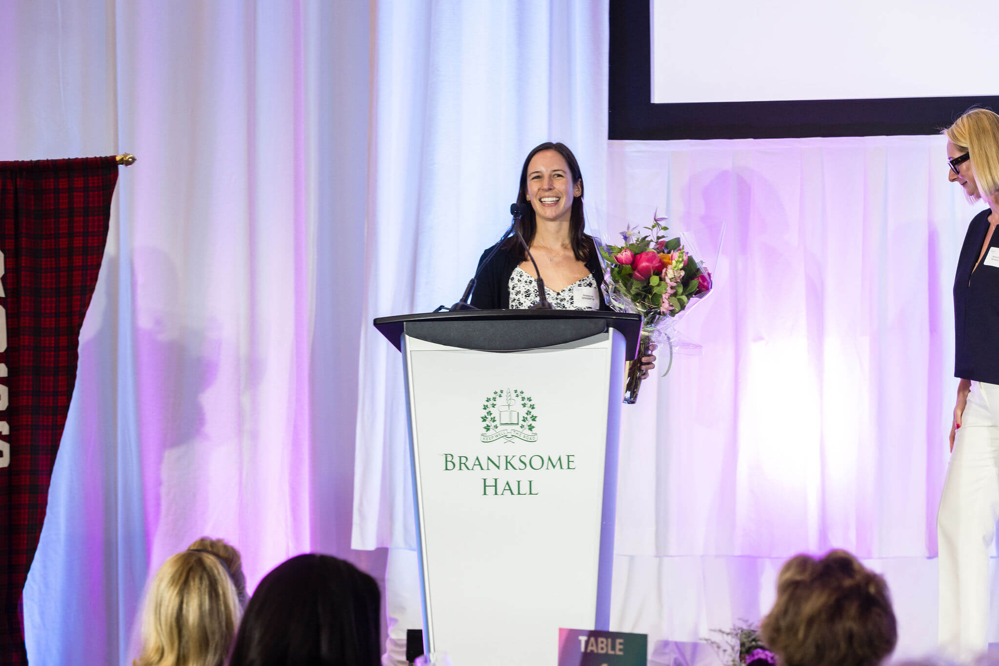 A woman holding a bouquet of flowers stands at a podium labeled "Branksome Hall," smiling as she addresses an audience, while another woman stands nearby on stage.