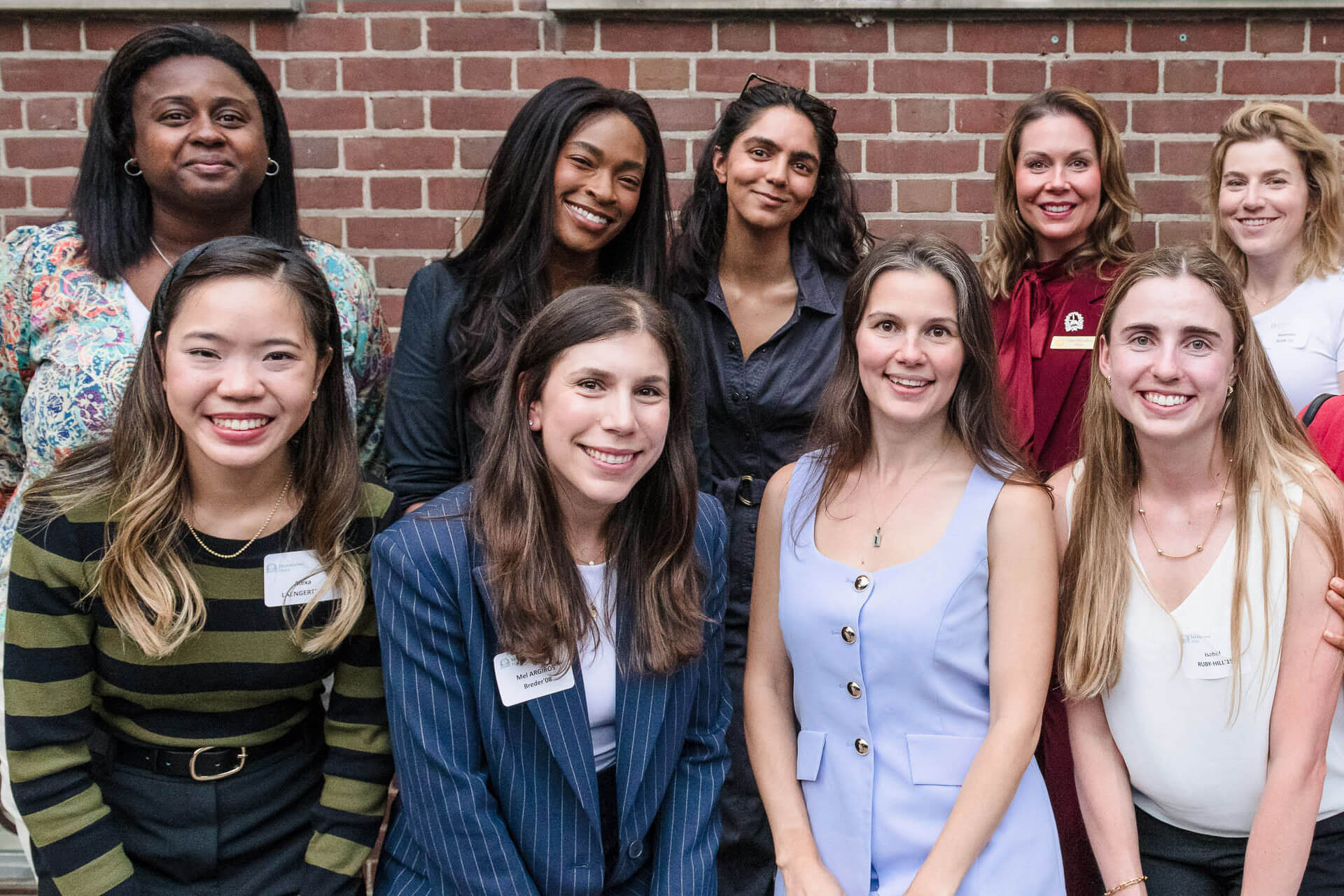 A group of nine women, smiling and standing close together, pose in front of a brick wall. They are wearing a variety of business casual outfits and name tags.