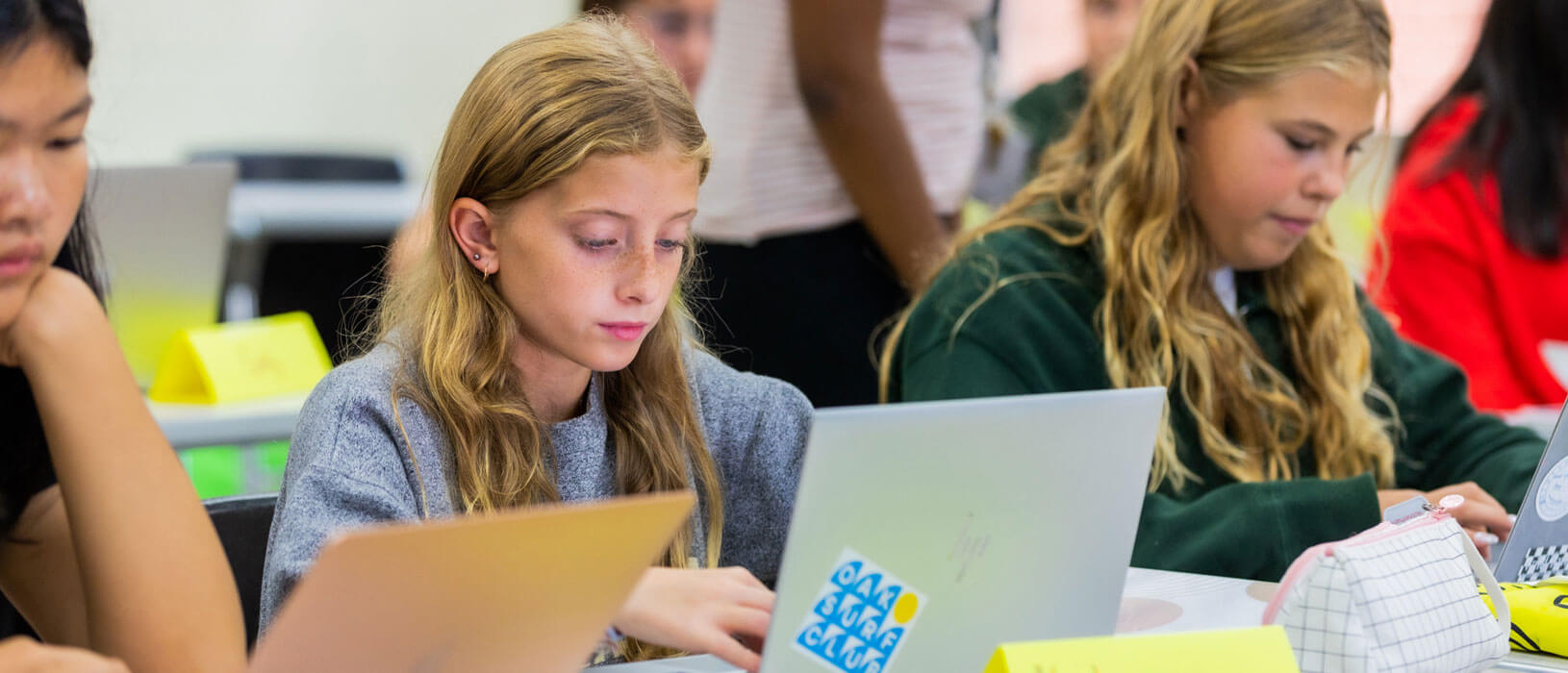 Three young students sit at desks using laptops in a classroom. The students are focused on their screens, with school supplies and name tags visible on the desks. A teacher stands in the background.