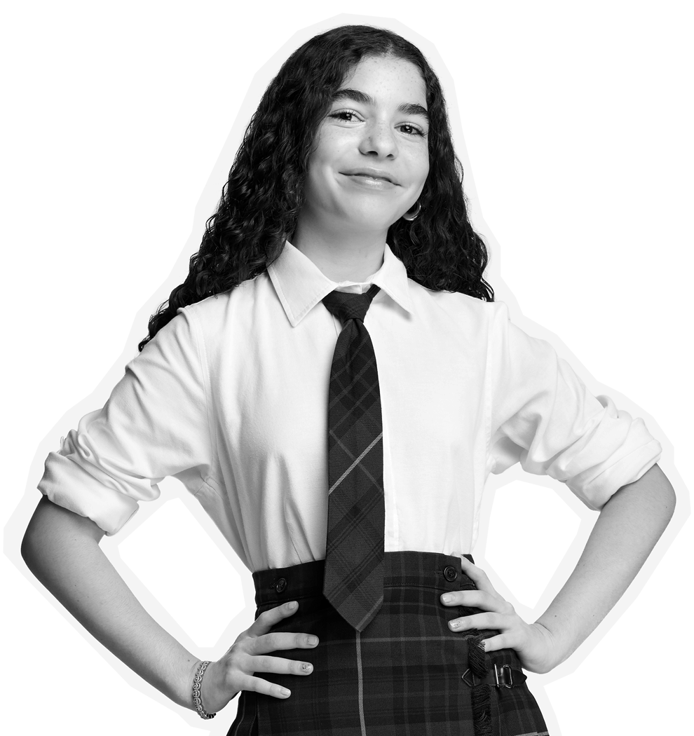 A confident young person with long curly hair smiles, standing with hands on hips. They wear a white shirt, plaid tie, and plaid skirt. The image is in black and white.
