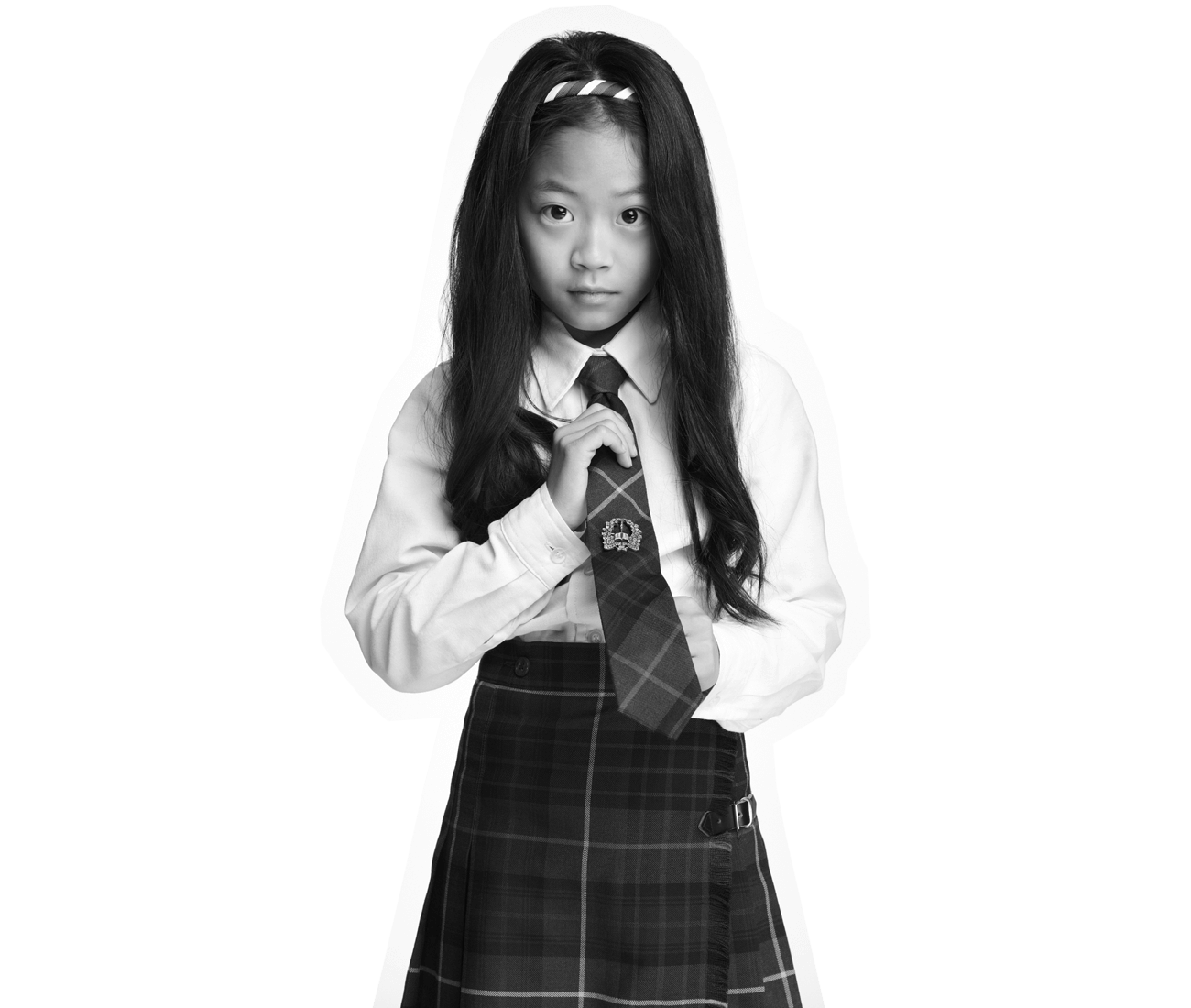 A young girl with long hair, wearing a plaid skirt, white shirt, and tie, stands facing forward and adjusting her tie. The image is in black and white.