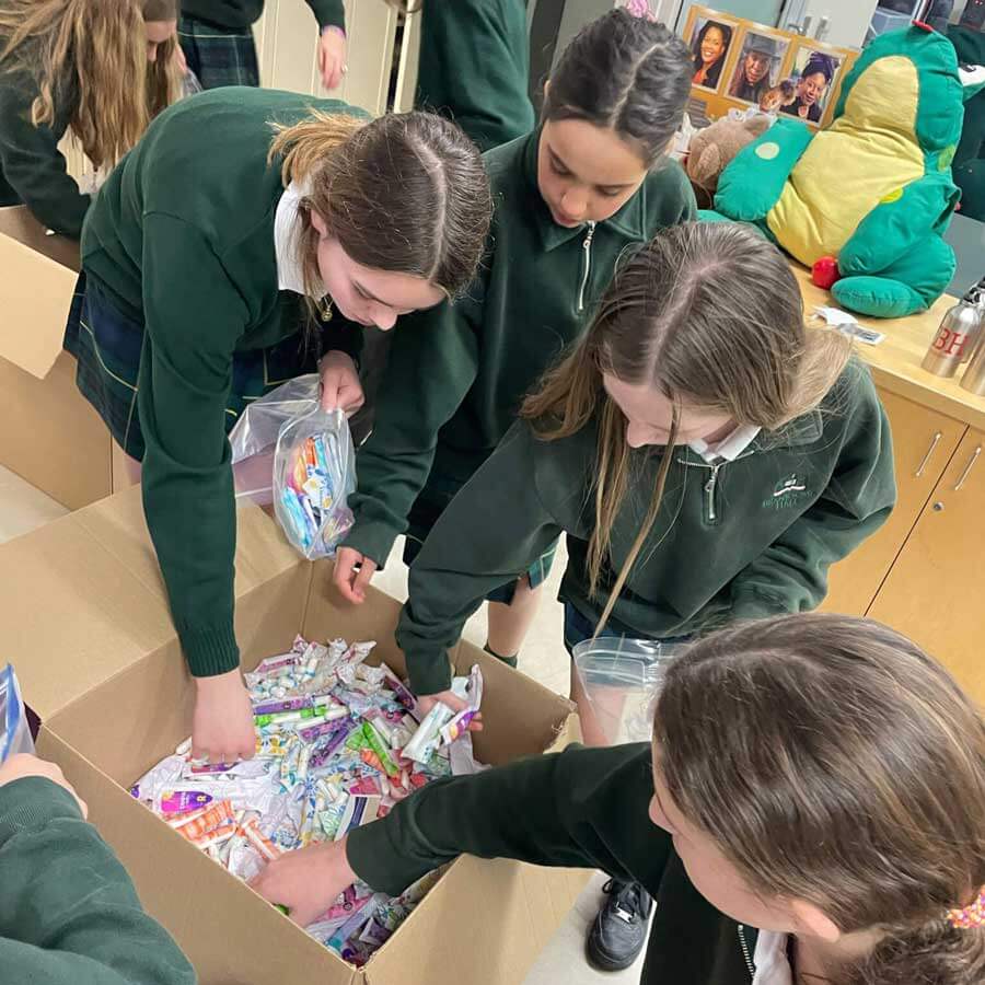 Four girls in green school uniforms are filling plastic bags with assorted toiletries or hygiene products from a large cardboard box on a table in a classroom.
