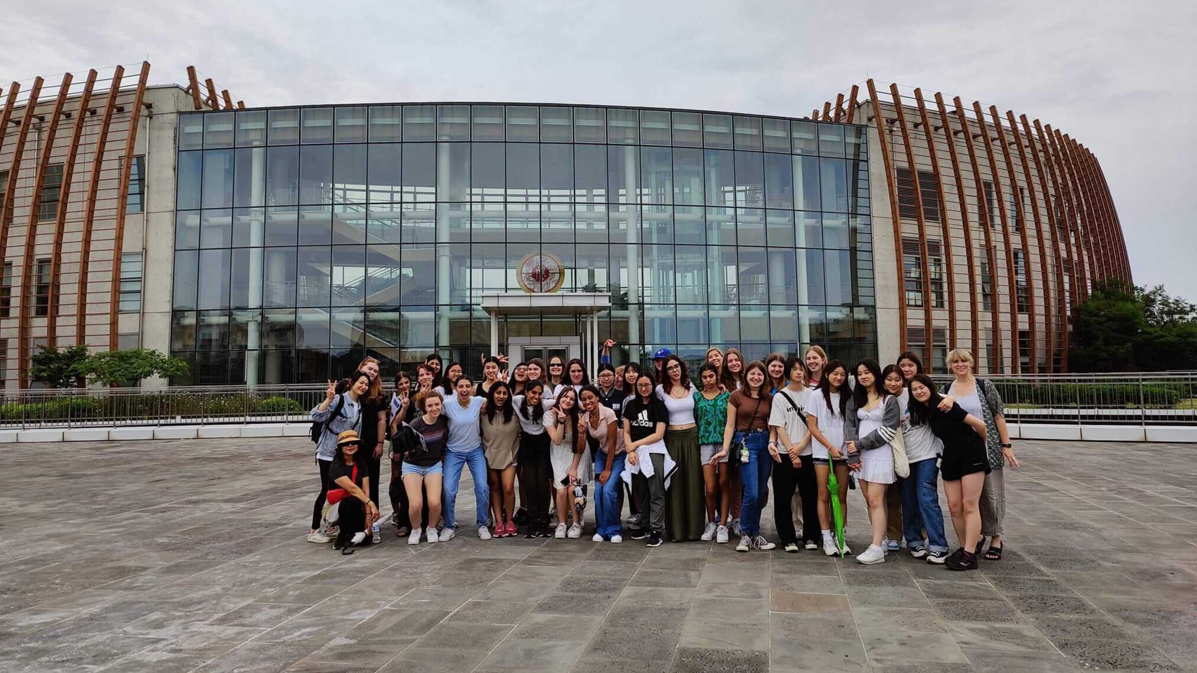 A large group of smiling young adults poses together outside a modern glass building with a curved wooden exterior on a cloudy day.