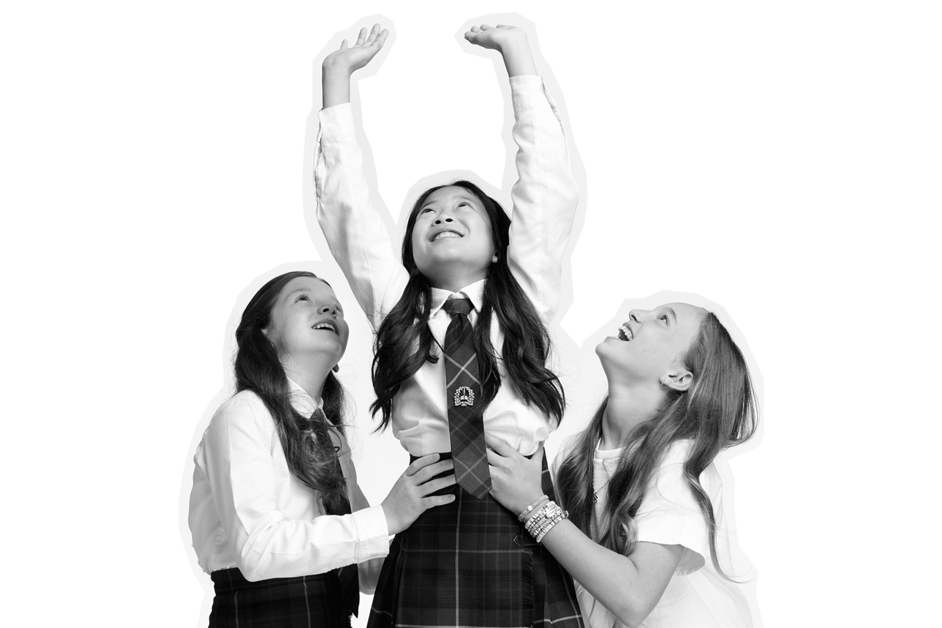 Three girls smile joyfully. One girl, in a school uniform, stands in the center, reaching both arms upward as the other two support her, looking up and laughing. The background is plain white.