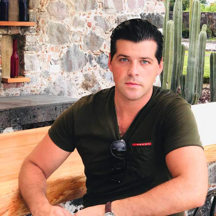 A man with dark slicked-back hair, wearing a dark green Prada t-shirt and sunglasses hanging from the collar, sits at a wooden table outdoors near stone walls and tall cacti.