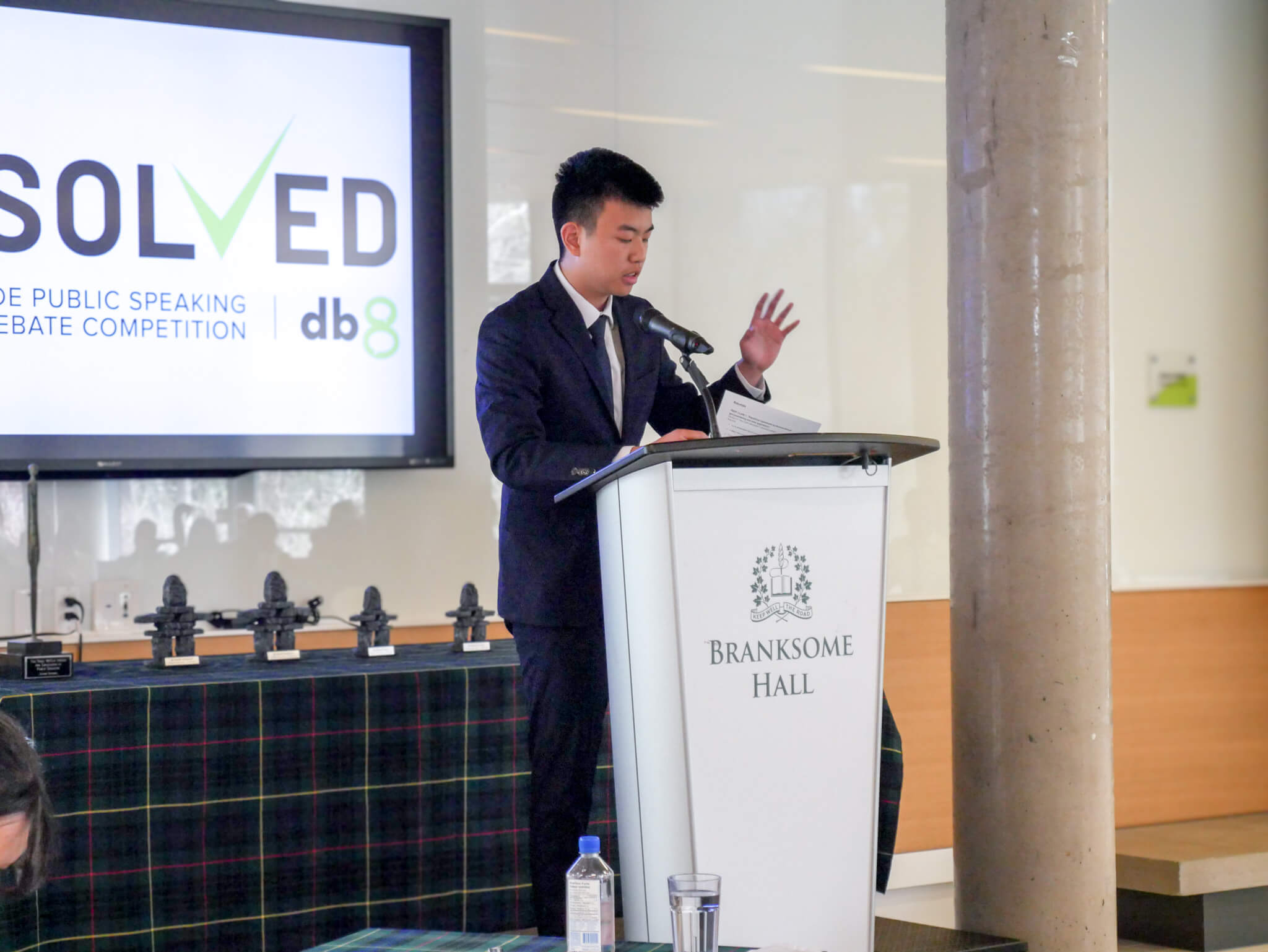 A young man in a suit speaks at a podium labeled "Branksome Hall" during a public speaking event. Behind him is a screen displaying the words "SOLVED" and several trophies are arranged on a table.