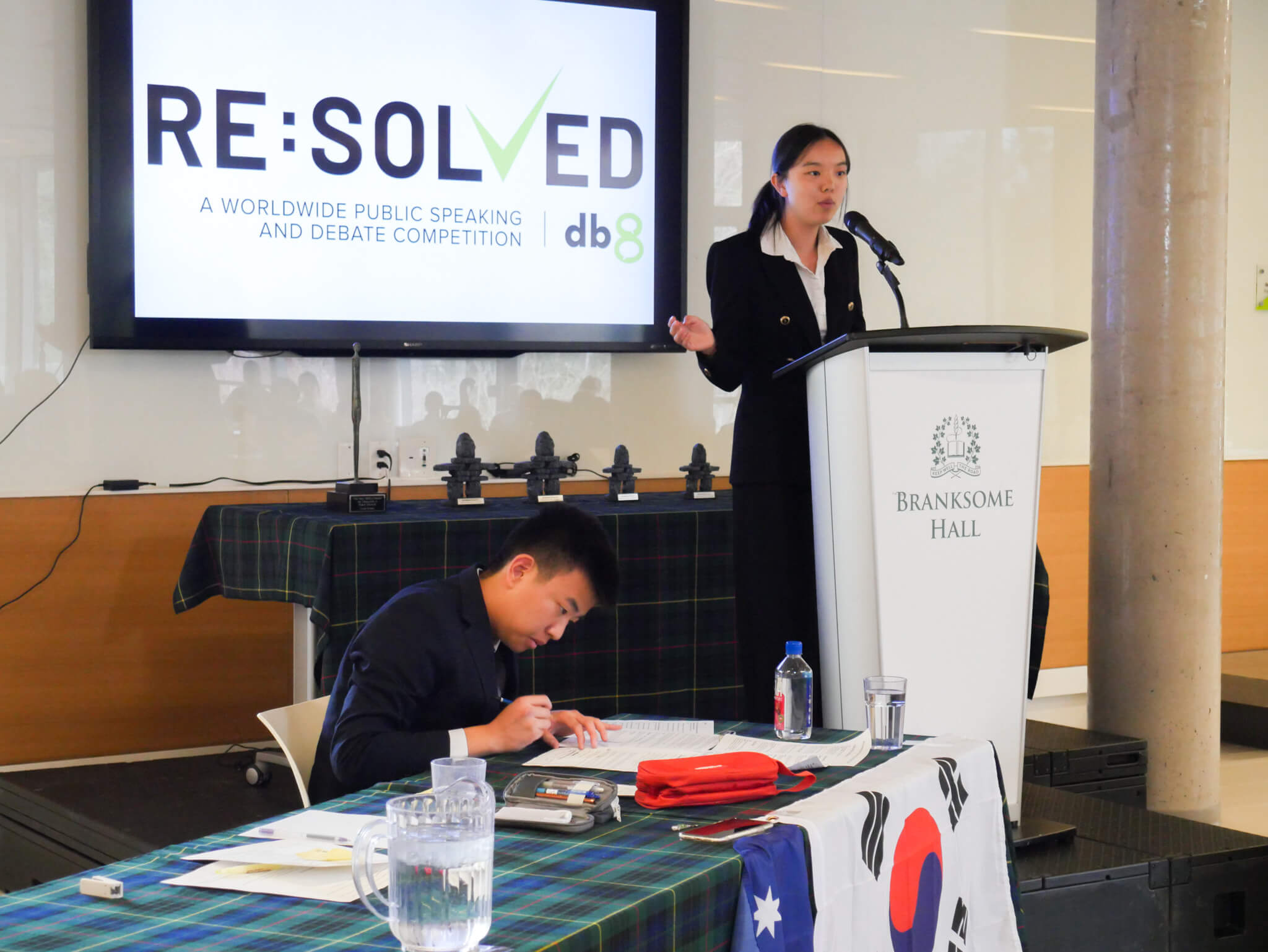 A young woman speaks at a podium labeled "Branksome Hall" during a public speaking and debate competition. A young man sits at a table in front, writing, with flags and a water pitcher beside him. A "RESOLVED" sign is displayed behind them.