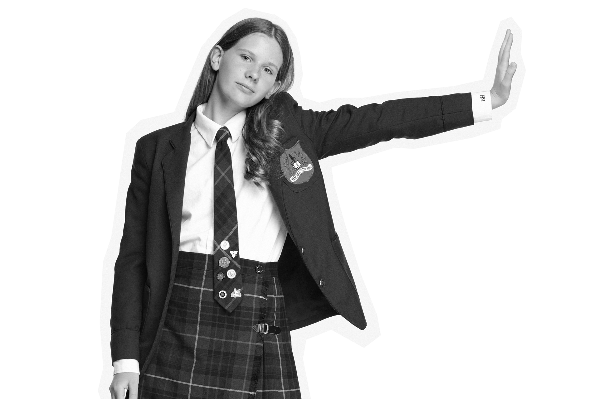 A girl in a school uniform stands with one arm extended sideways. She wears a blazer with a crest, a tie, and a plaid skirt. The image is in black and white.