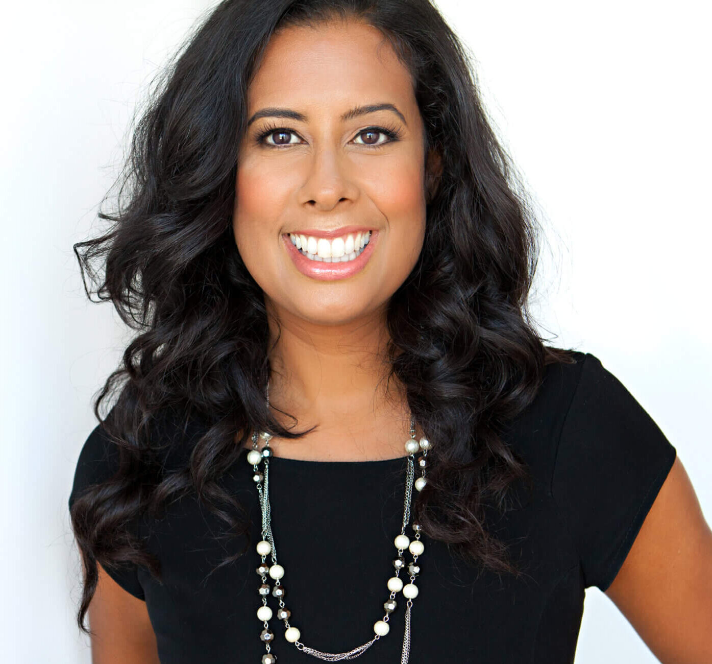 A woman with long, wavy dark hair wearing a black dress and a long beaded necklace is smiling at the camera in front of a plain white background.