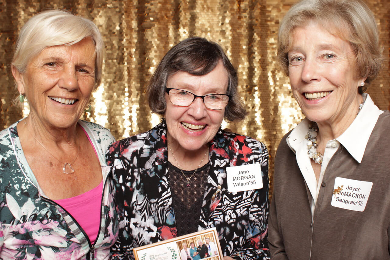 Three smiling older women stand together in front of a gold sequin backdrop, each wearing a name tag and casual clothing. The woman in the center holds a printed photograph.