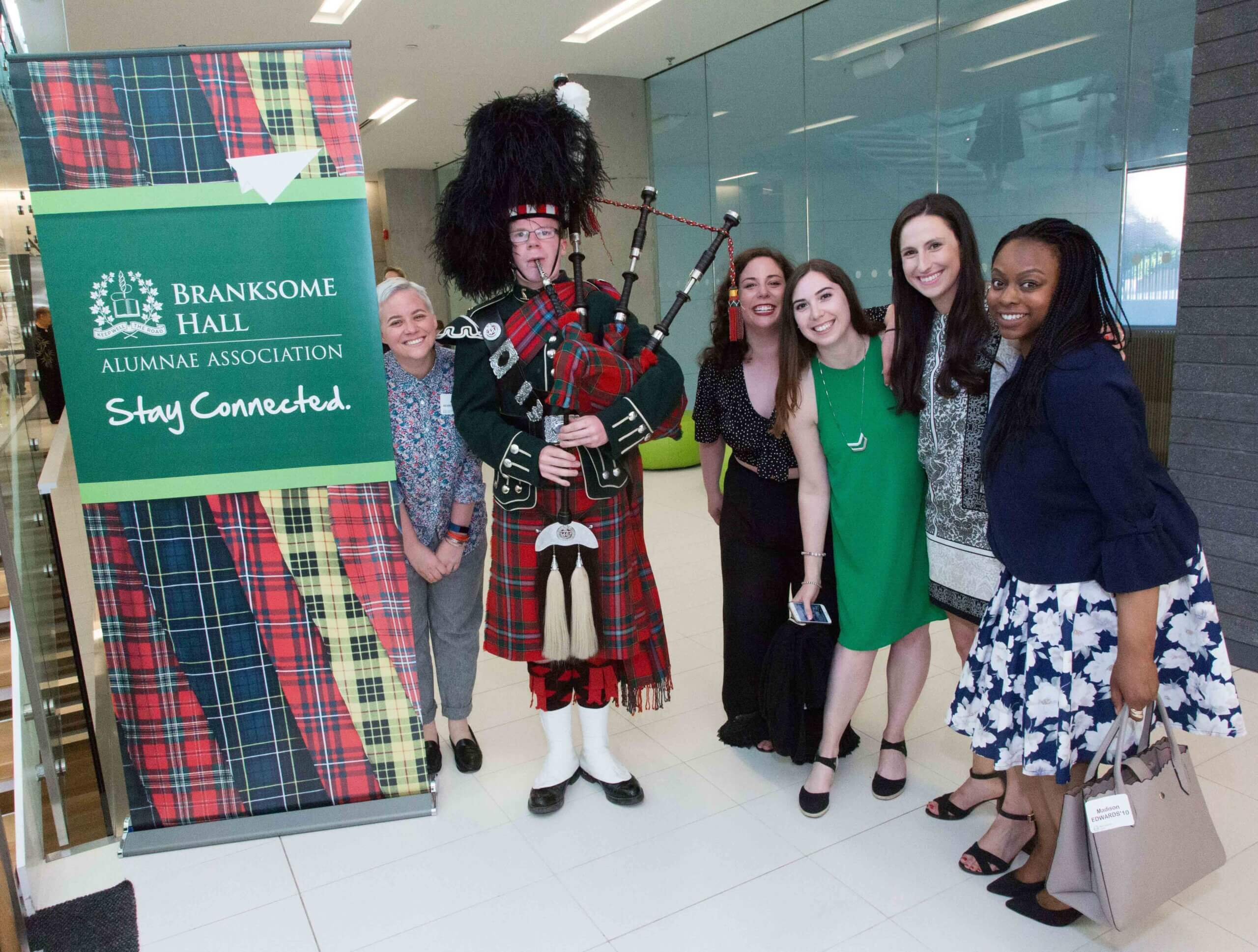 Six people pose indoors beside a “Branksome Hall Alumnae Association” sign. Five women stand smiling next to a bagpiper in traditional Scottish attire. The setting is bright and modern.