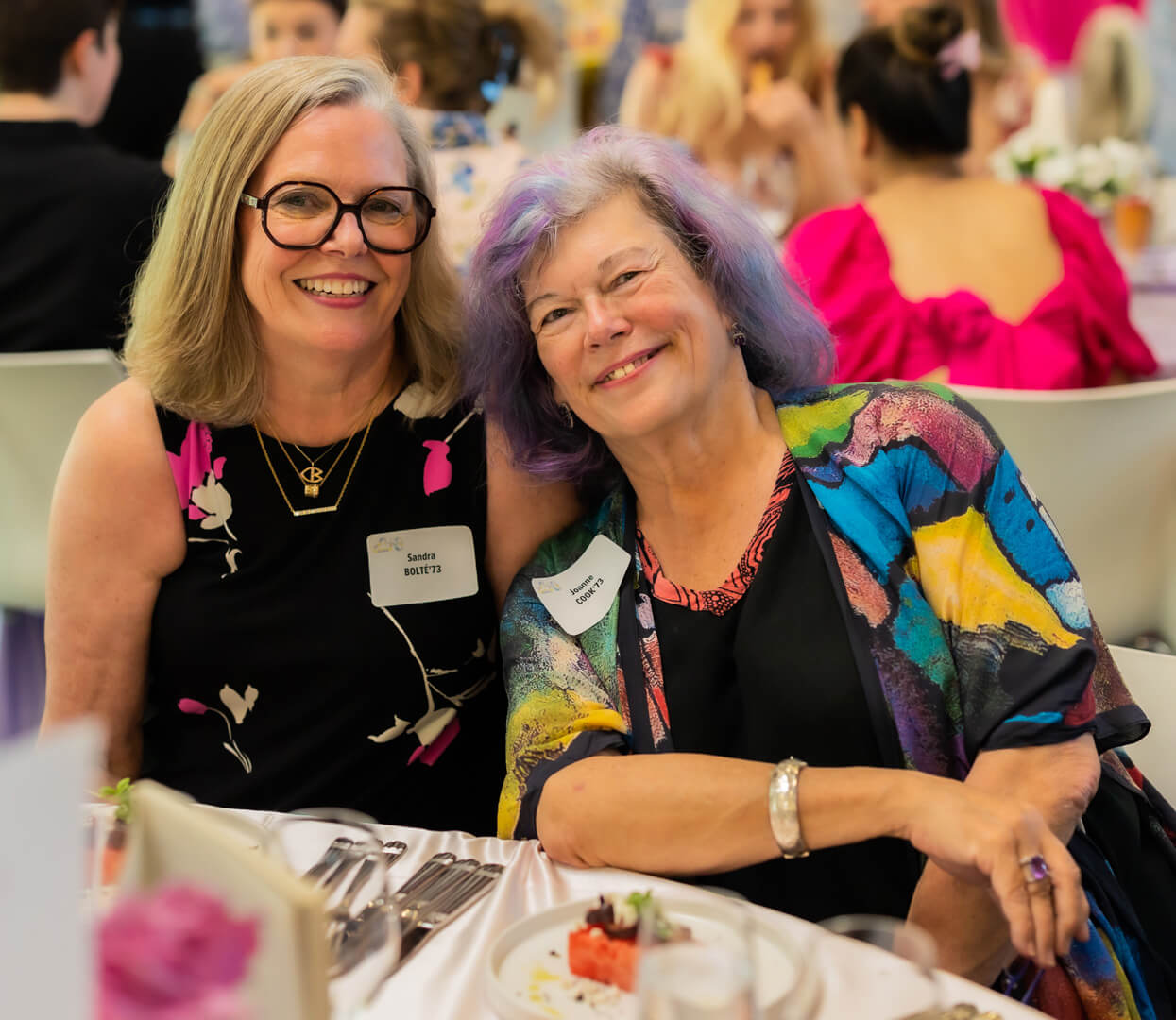 Two smiling women sit close together at a table during a social event. Both wear name tags and colorful outfits. Table settings and dessert plates are in front of them, with other people blurred in the background.