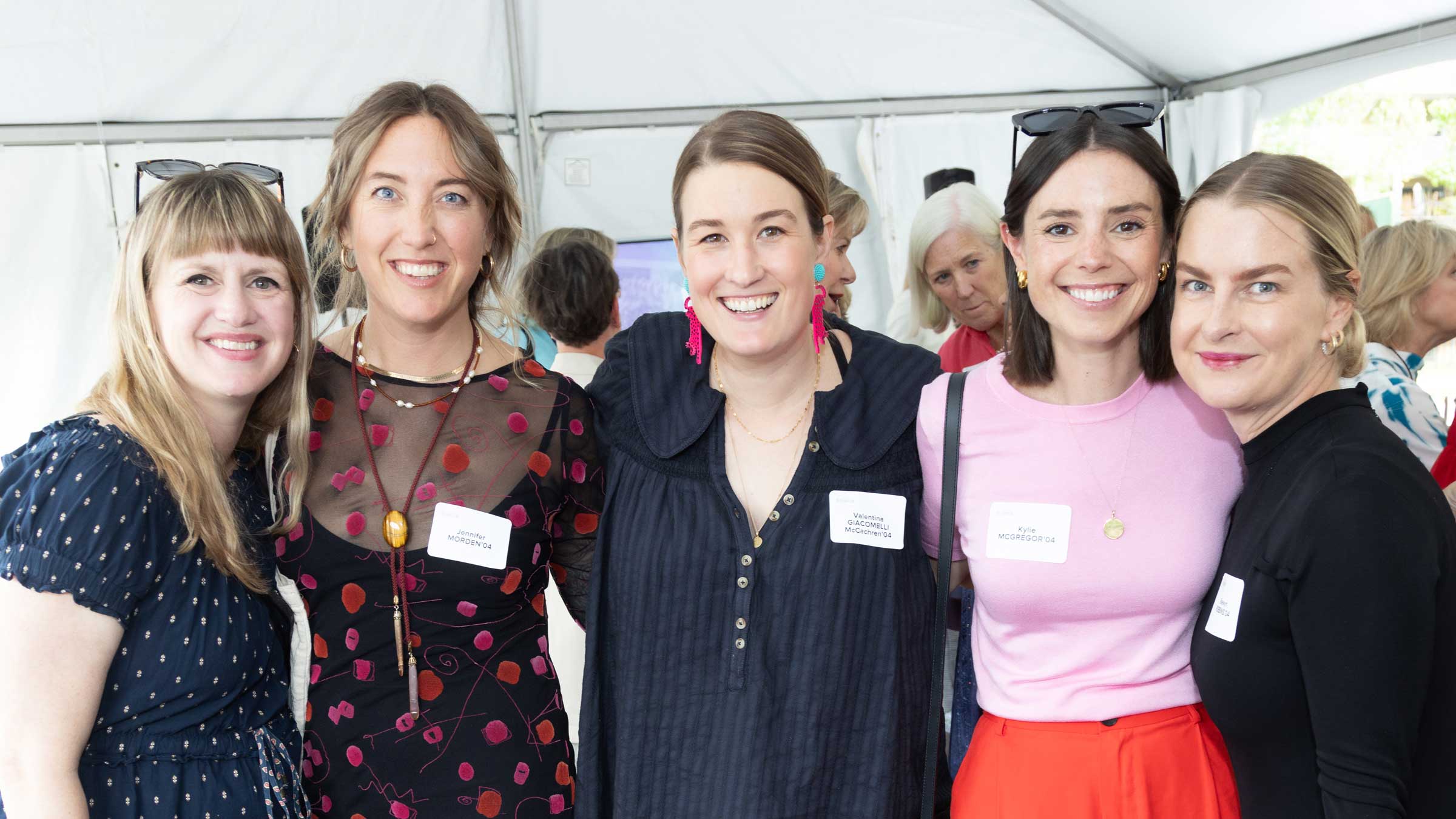 Five women smile and pose together at an indoor event, each wearing name tags. Others are visible in the background, and they appear to be gathered under a white tent.