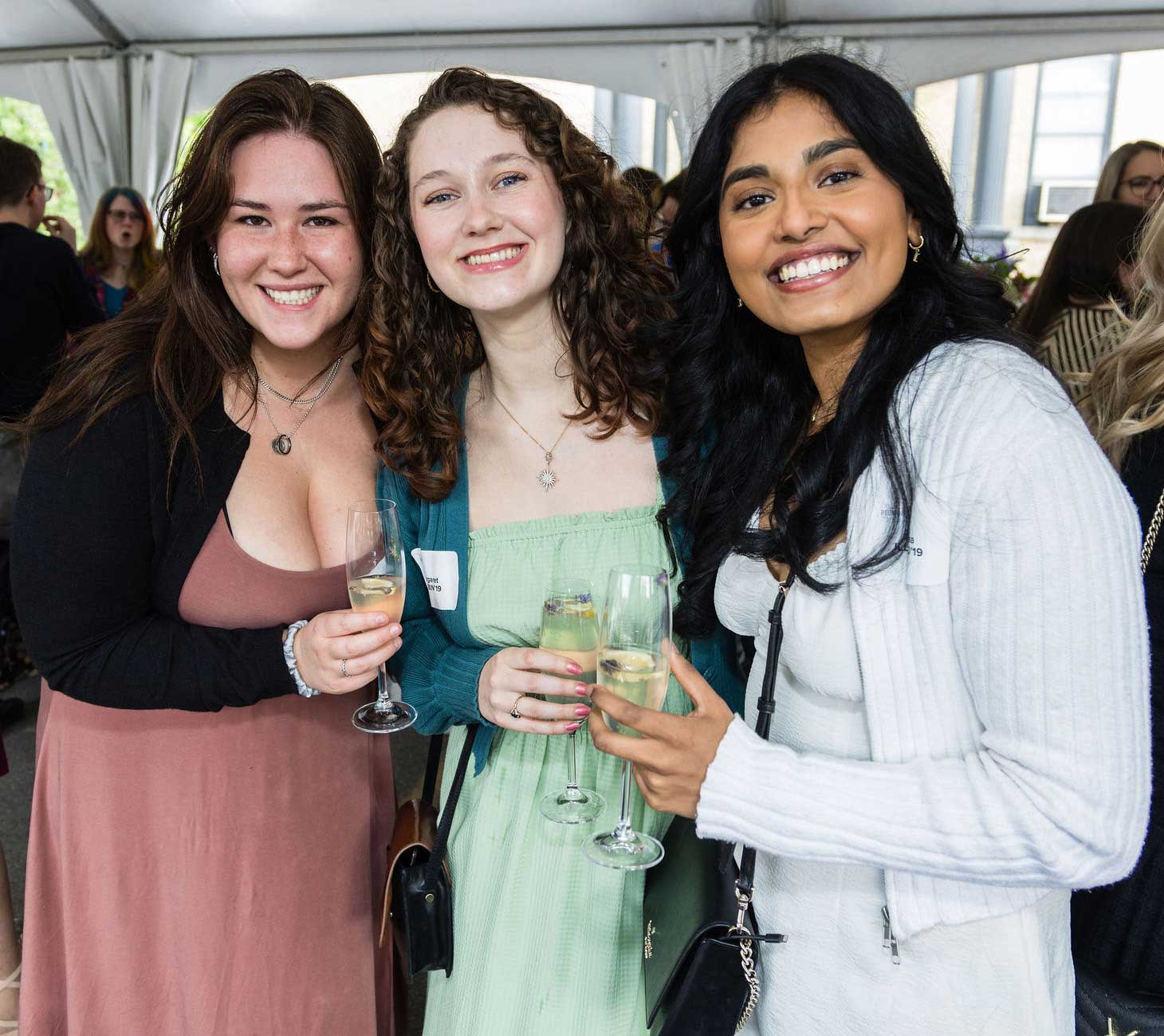 Three women stand close together, smiling and holding glasses of champagne at an outdoor event under a tent. Other people are visible in the background, suggesting a lively social gathering.