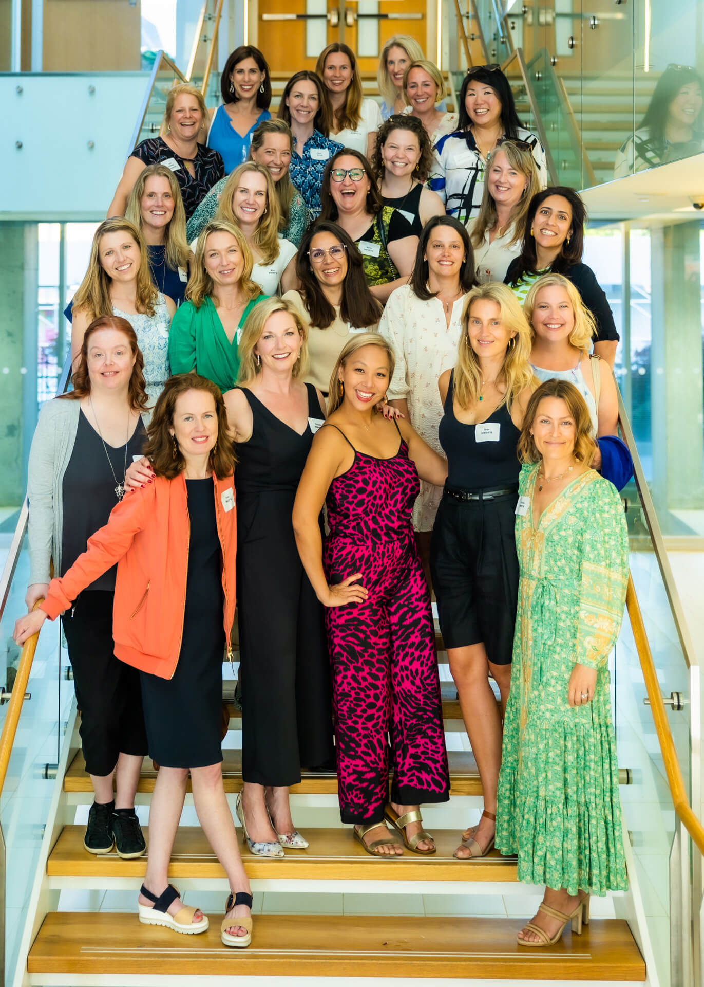 A group of women smiling and posing together on a staircase in a brightly lit, modern building with glass railings and wood accents. Most are wearing summer dresses or casual clothes.