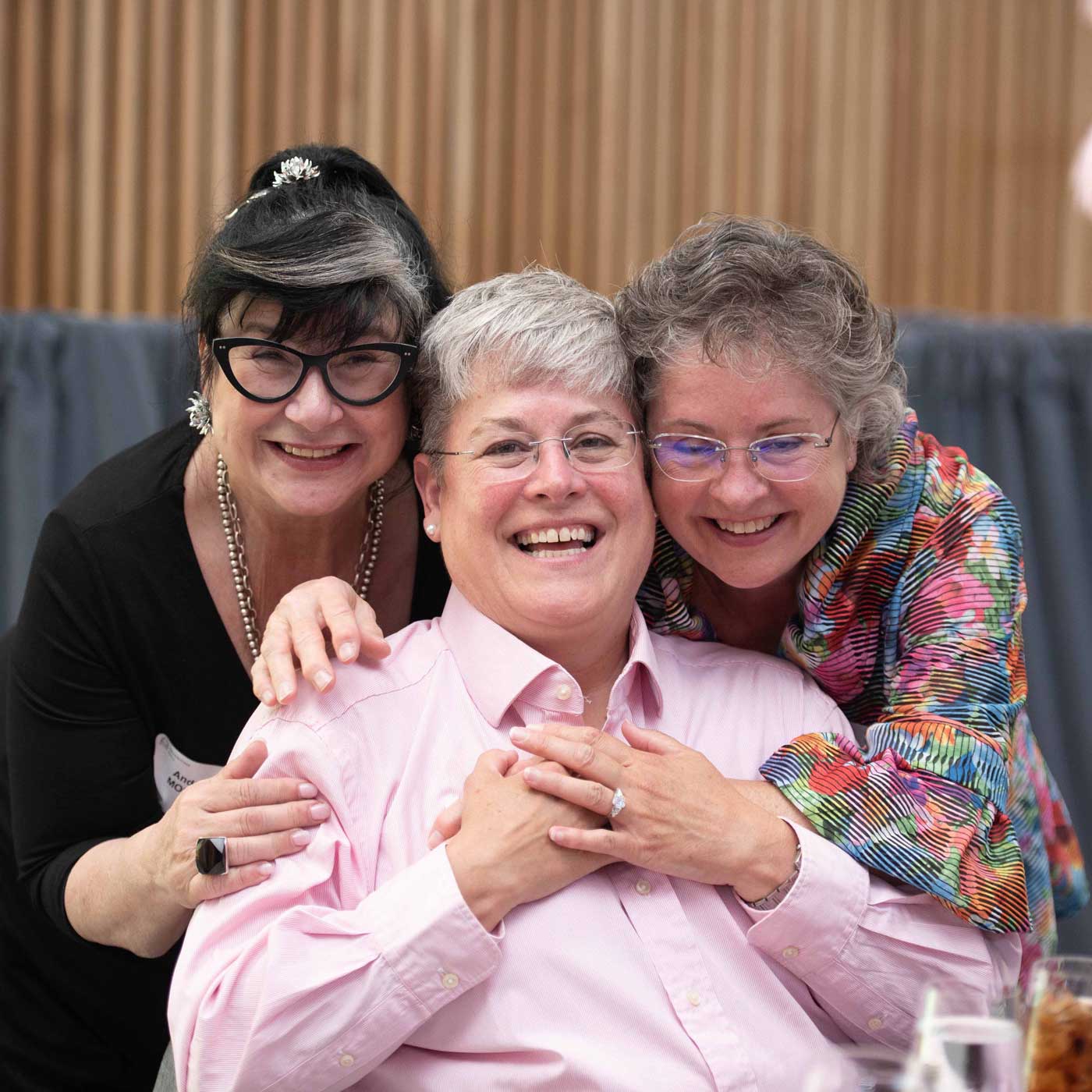 Three smiling older women pose together indoors. The woman in the middle wears glasses and a pink shirt, while the two women on either side hug her warmly and lean in close, all looking joyful and happy.