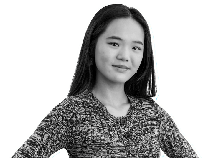 A young woman with long dark hair, wearing a textured patterned sweater, stands confidently with a slight smile and her hands on her hips. The image is in black and white with a white outline around her.