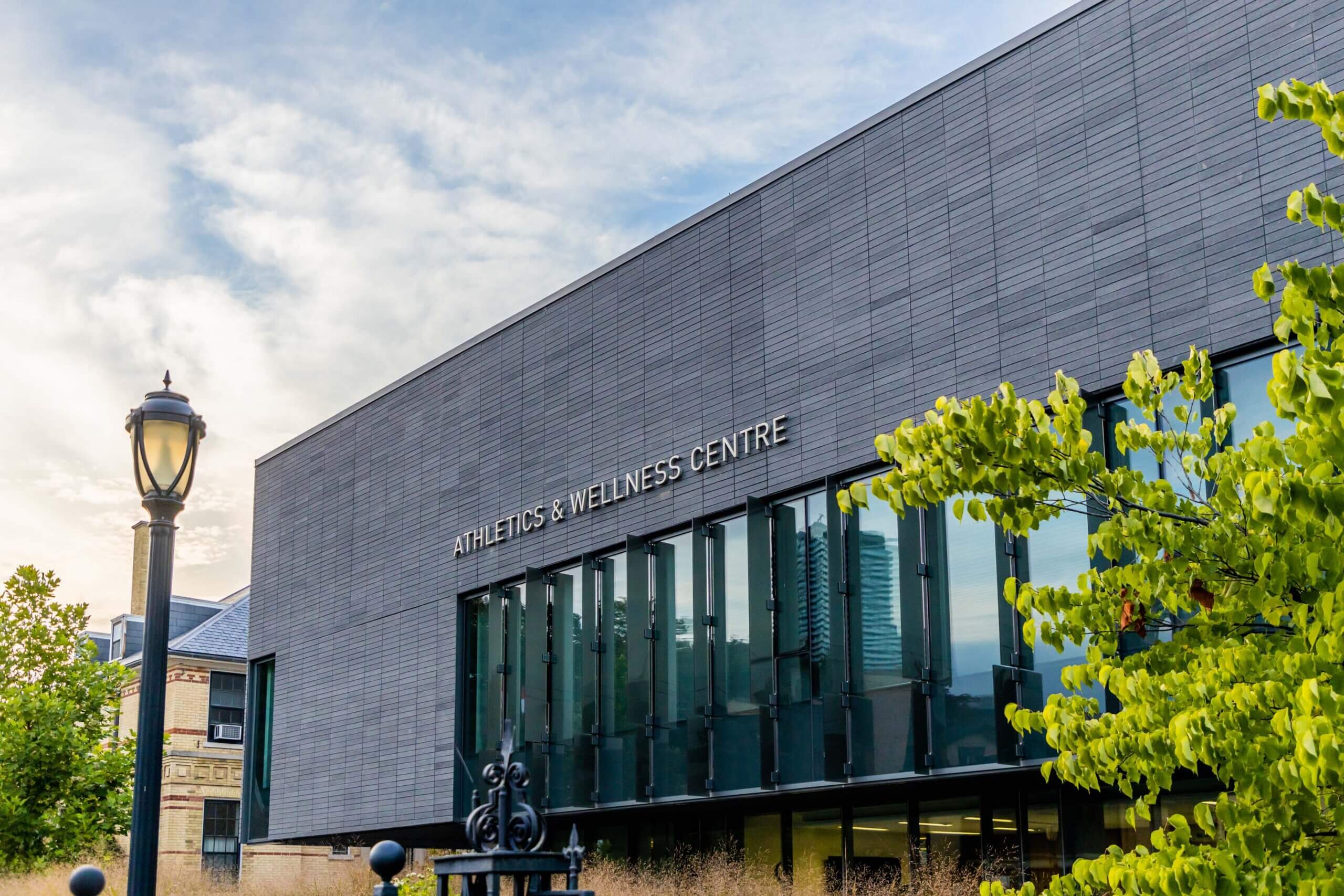 A modern building labeled "Athletics & Wellness Centre" with large windows, a streetlamp, and green trees in the foreground under a partly cloudy sky.