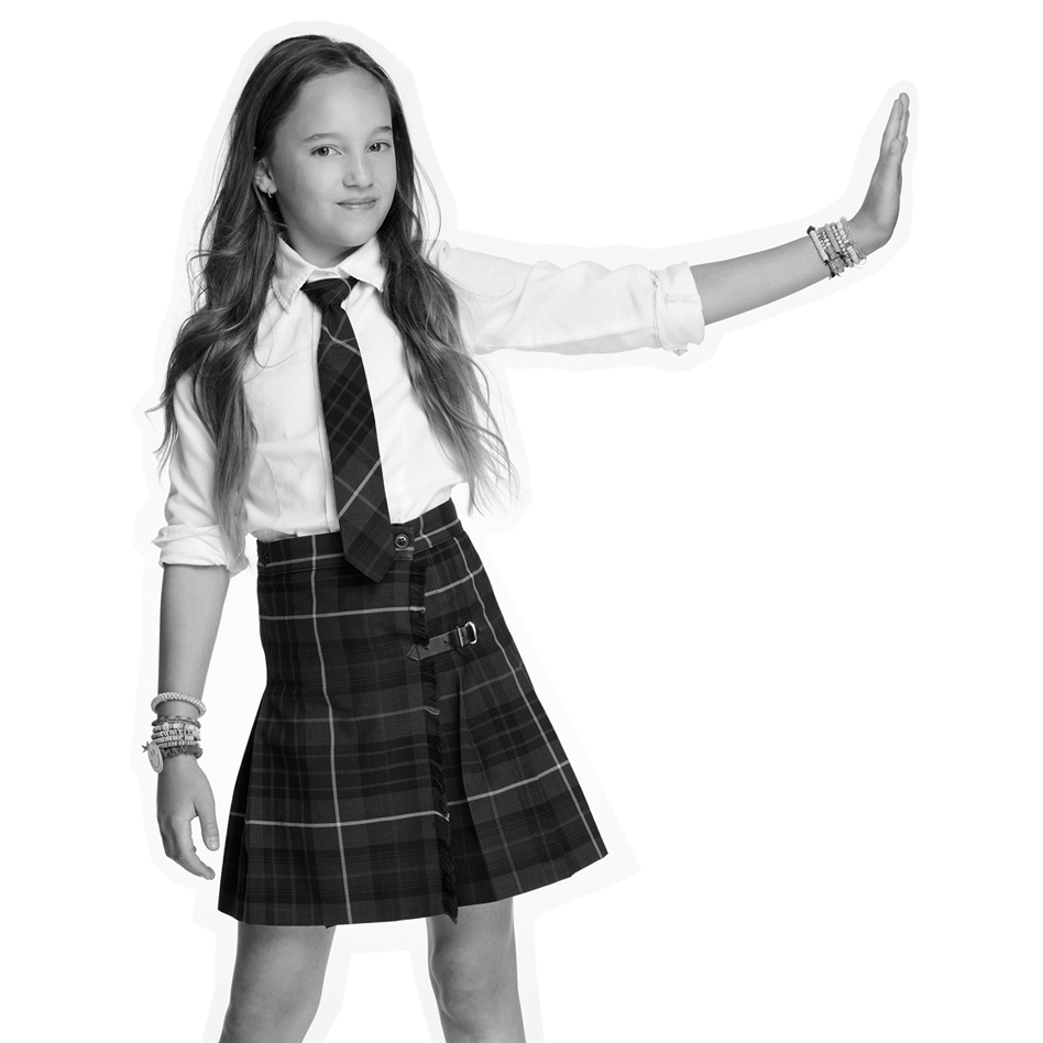 A young girl wearing a white shirt, plaid skirt, and tie stands confidently with one arm extended to the side, palm facing outward. She has long hair and wears several bracelets. The image is in black and white.