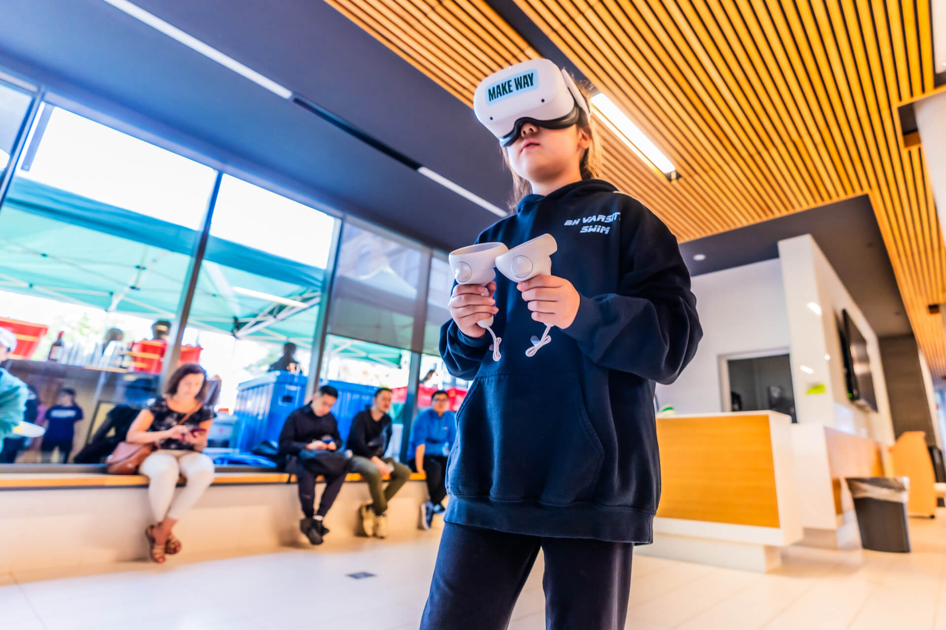 A child wearing a VR headset and holding controllers stands indoors, immersed in virtual reality. Several people are seated in the background near large windows and a wooden ceiling.