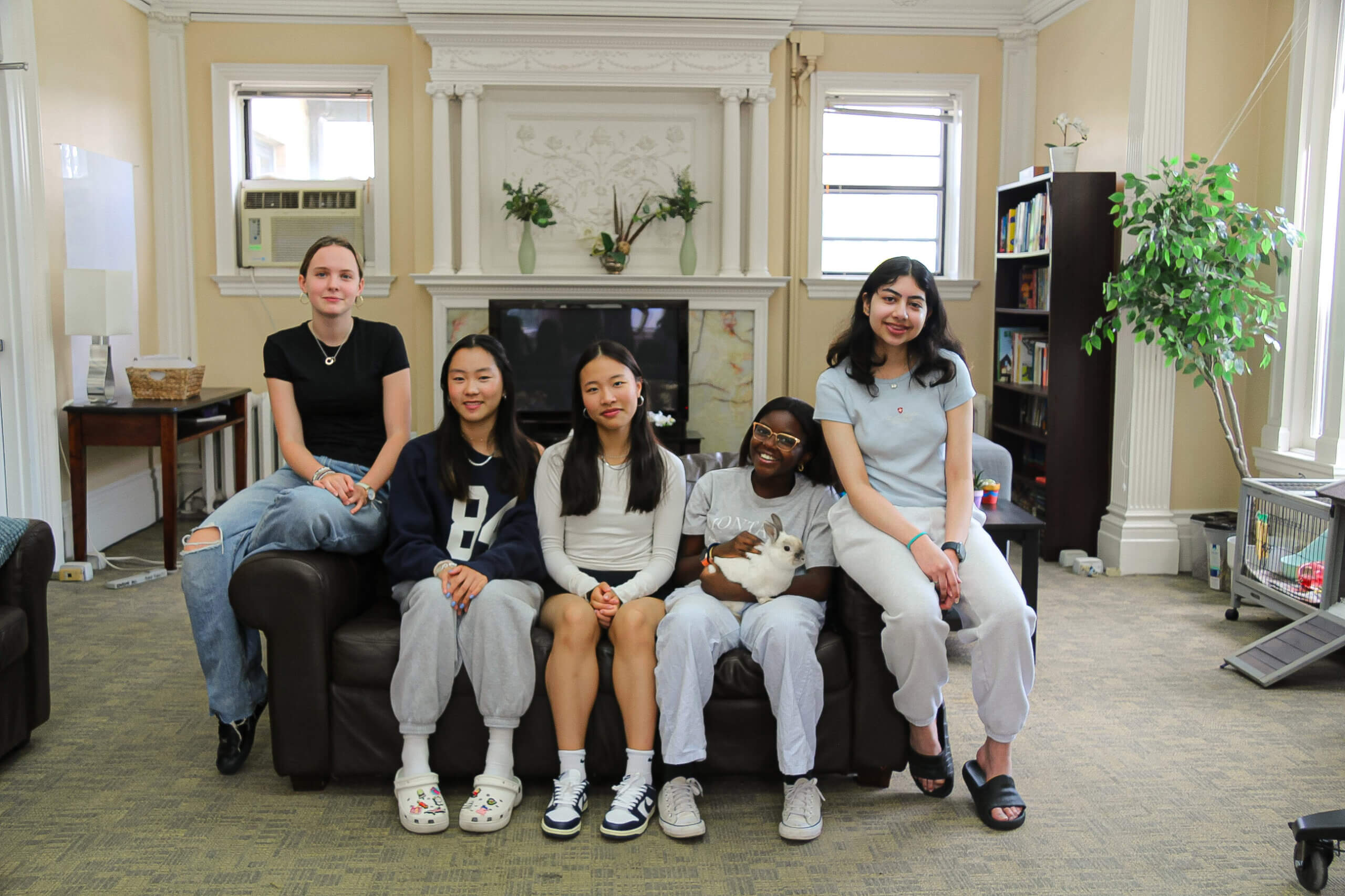 Five young women sit on a dark sofa in a bright room with large windows, a fireplace, bookshelves, and a potted plant. One person is holding a white cat, and they are all smiling at the camera.