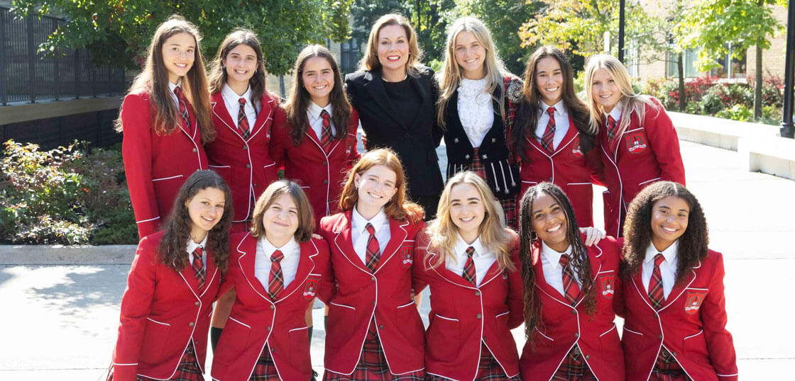 A group of thirteen smiling young women, most wearing matching red blazers and plaid skirts, pose outdoors in the sunshine with two adults standing behind them.
