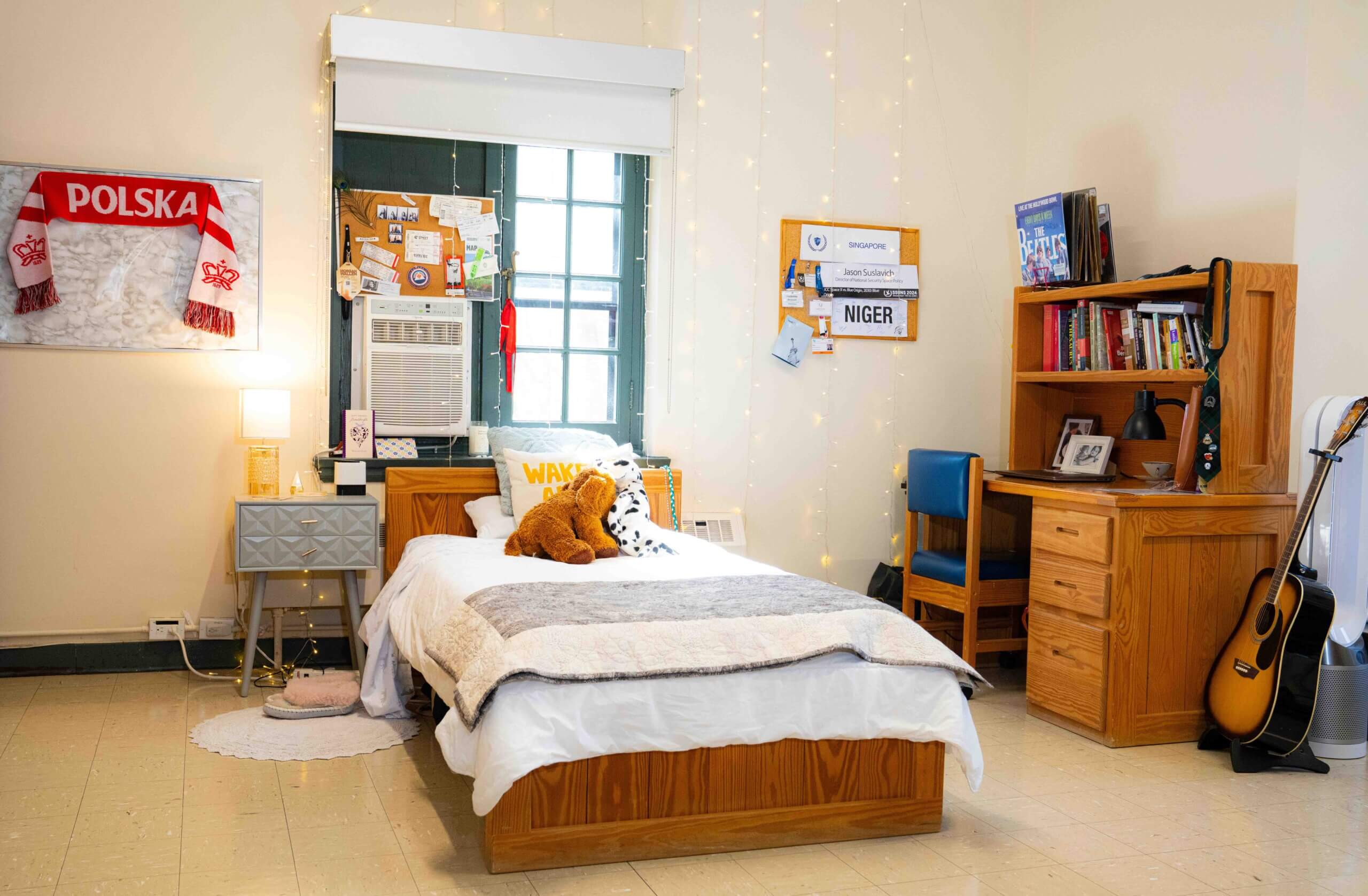 A cozy dorm room with a bed, stuffed toy, and string lights, a desk with books and a guitar, a wall-mounted air conditioner, and decorations including a Polska scarf and a corkboard with notes.