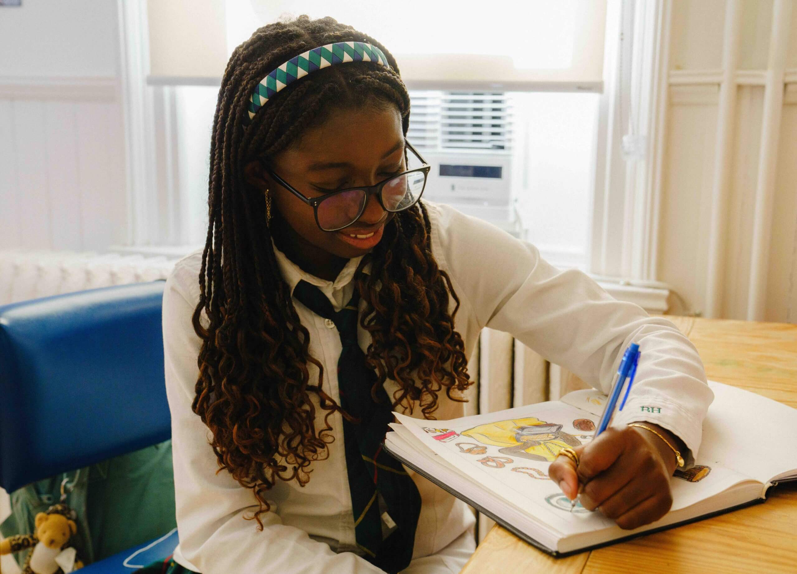 A girl with glasses and braided hair sits at a table, smiling as she colors in a drawing on a sheet of paper with a blue pen. She wears a white shirt and a tie, with a headband in her hair.