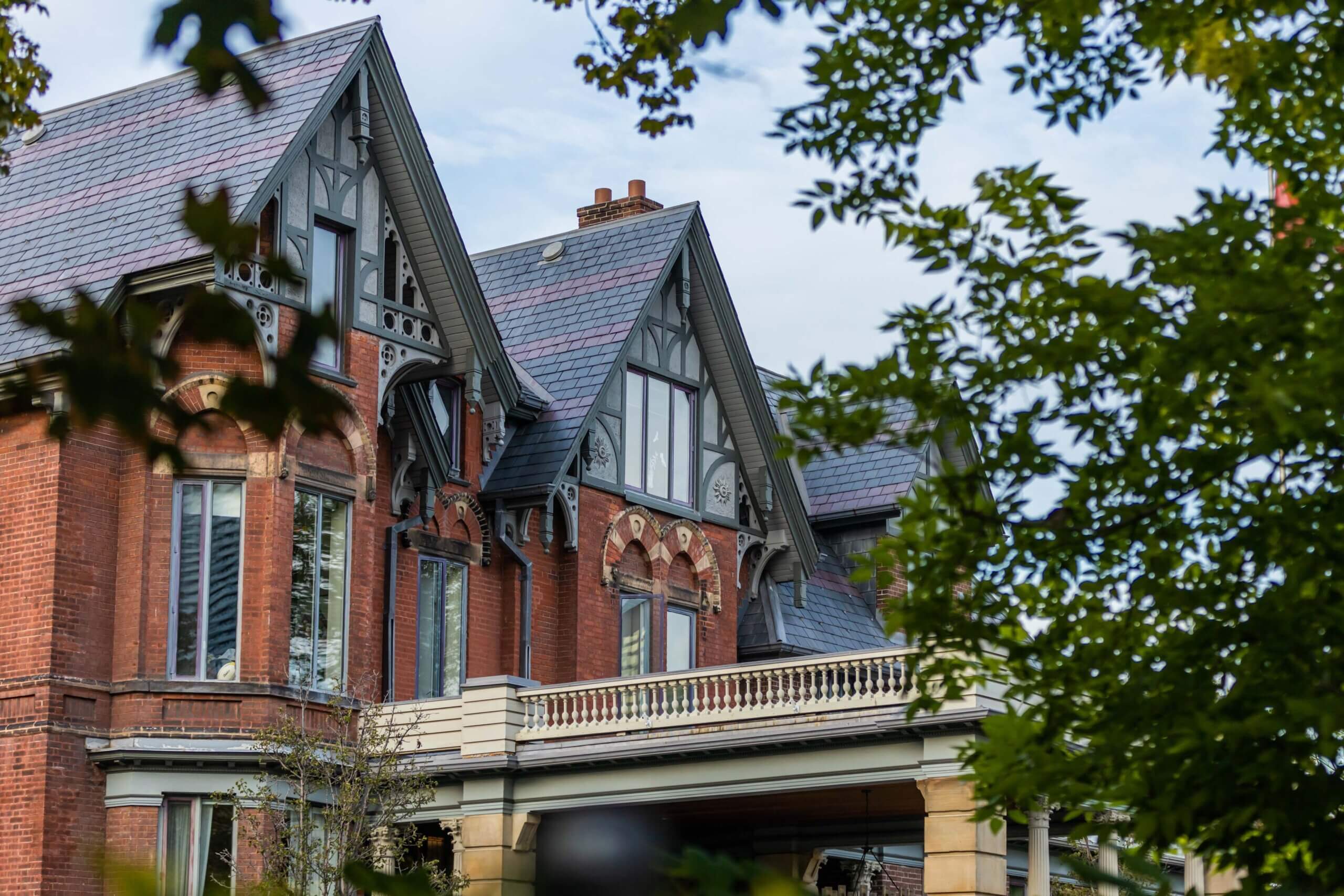 A large, historic red brick house with ornate gables, tall windows, and a balcony with a decorative railing, partially framed by green leafy branches.