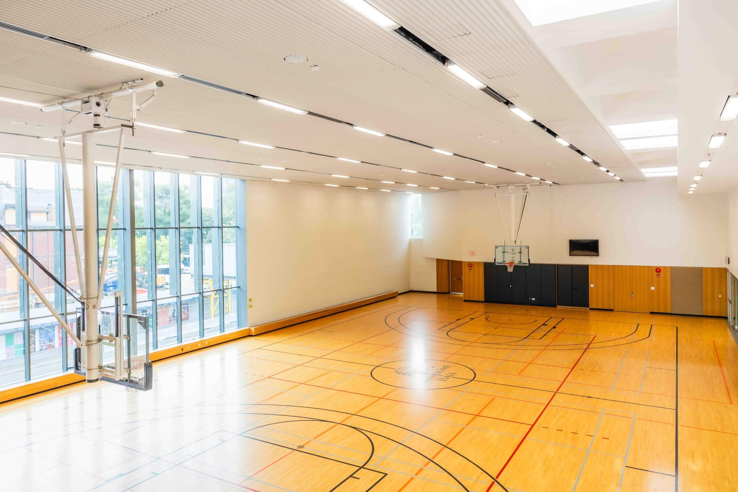 A spacious, empty indoor basketball court with wooden floors, large windows along one wall, and basketball hoops on either end. The court is well-lit with natural and artificial light.