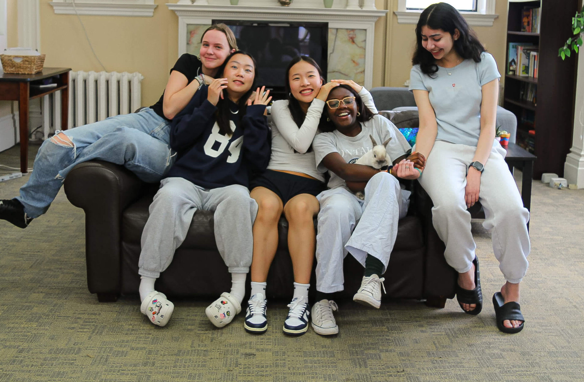Five young women sit closely together on a couch in a cozy, well-lit living room, smiling and laughing. They appear relaxed and happy, dressed casually in comfortable clothes and slippers.