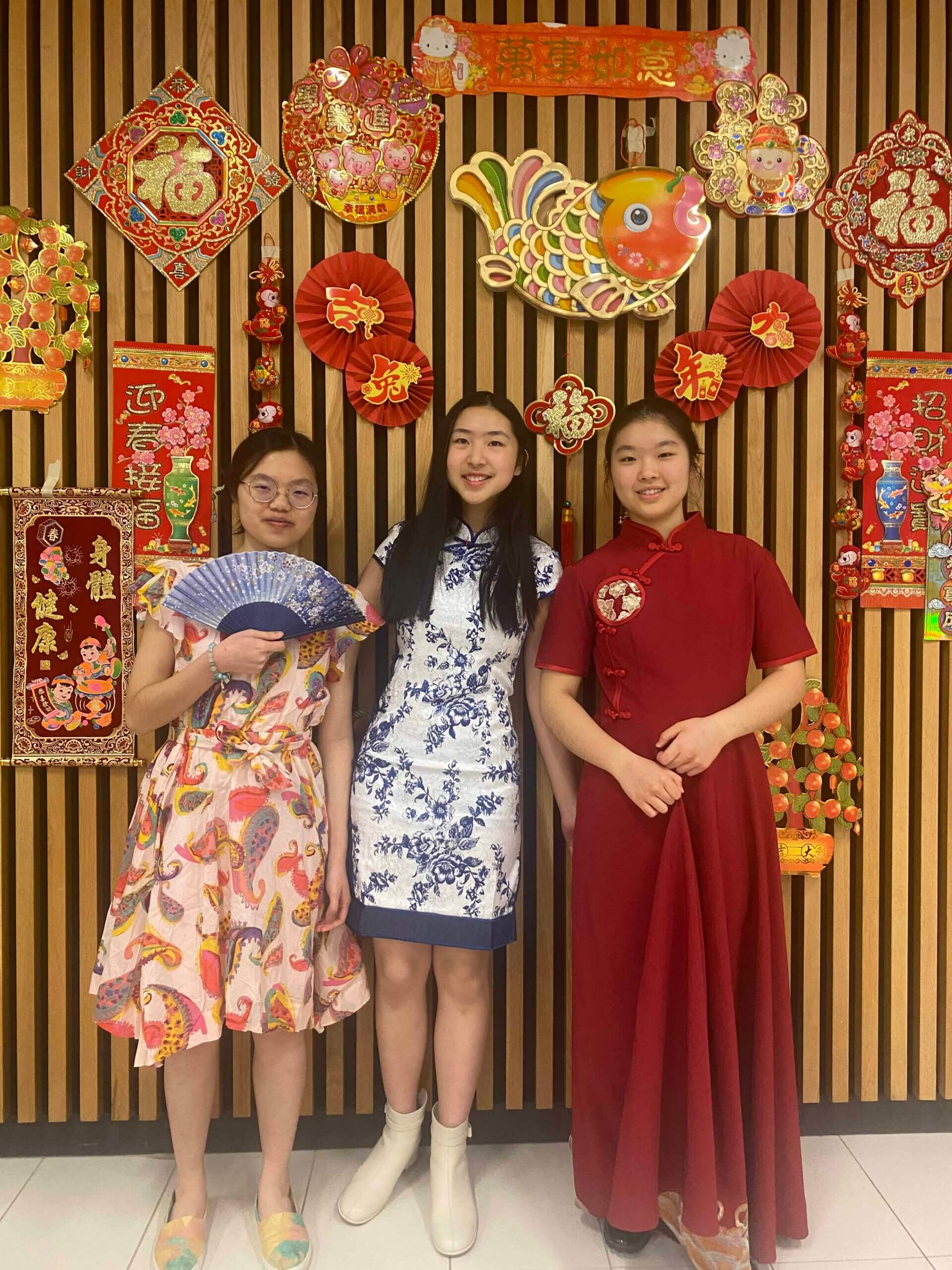 Three young women in traditional and festive dresses stand in front of a wall decorated with vibrant red and gold Chinese New Year ornaments, including a fish, fans, and banners.