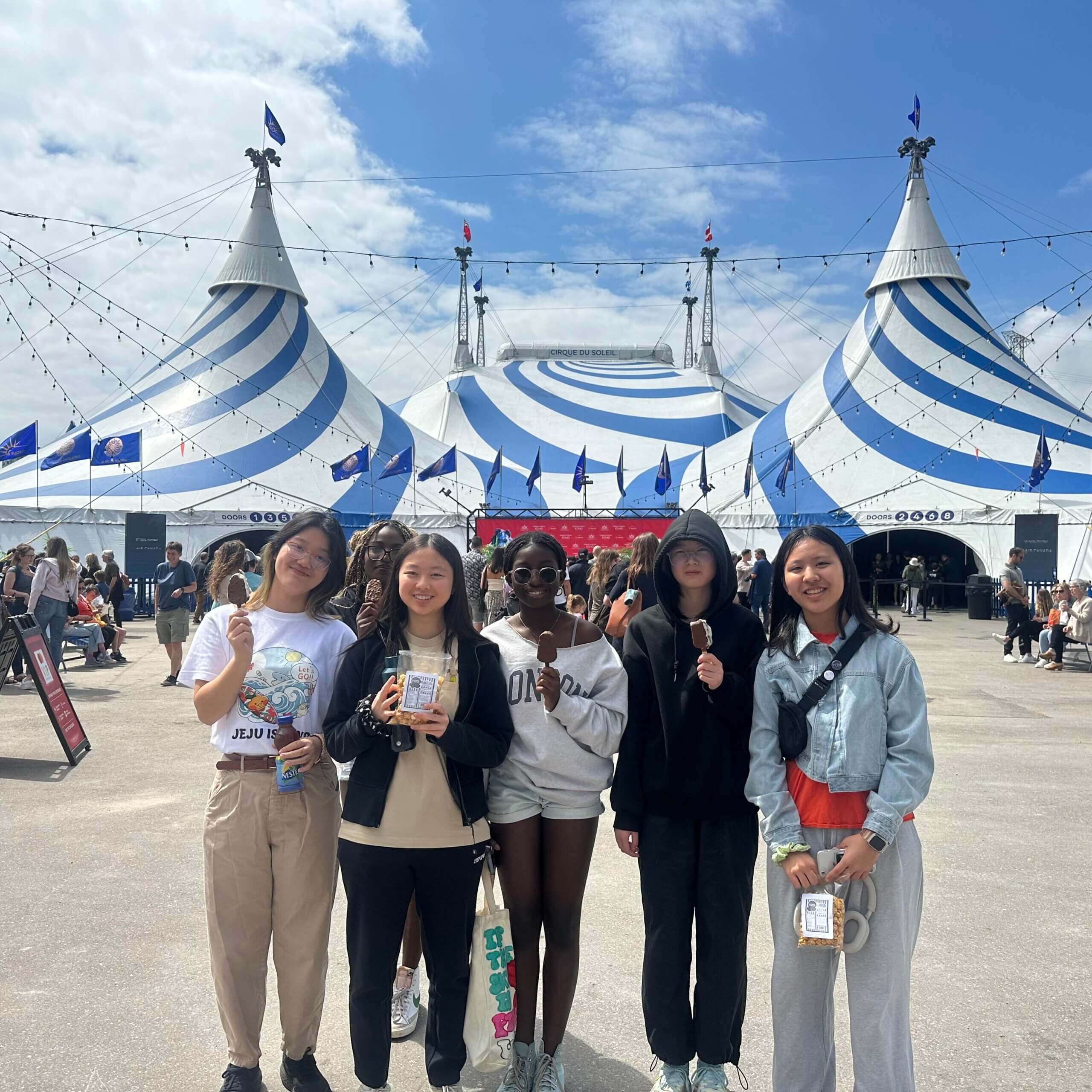 Five young people stand smiling in front of a large blue and white striped circus tent on a sunny day. There are crowds and string lights in the background, and everyone appears excited.