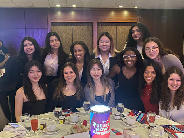 A group of young women dressed in semi-formal attire smile for a photo around a banquet table with drinks, plates, and a "Future Now Awards" centerpiece. The background shows a wood-paneled wall.