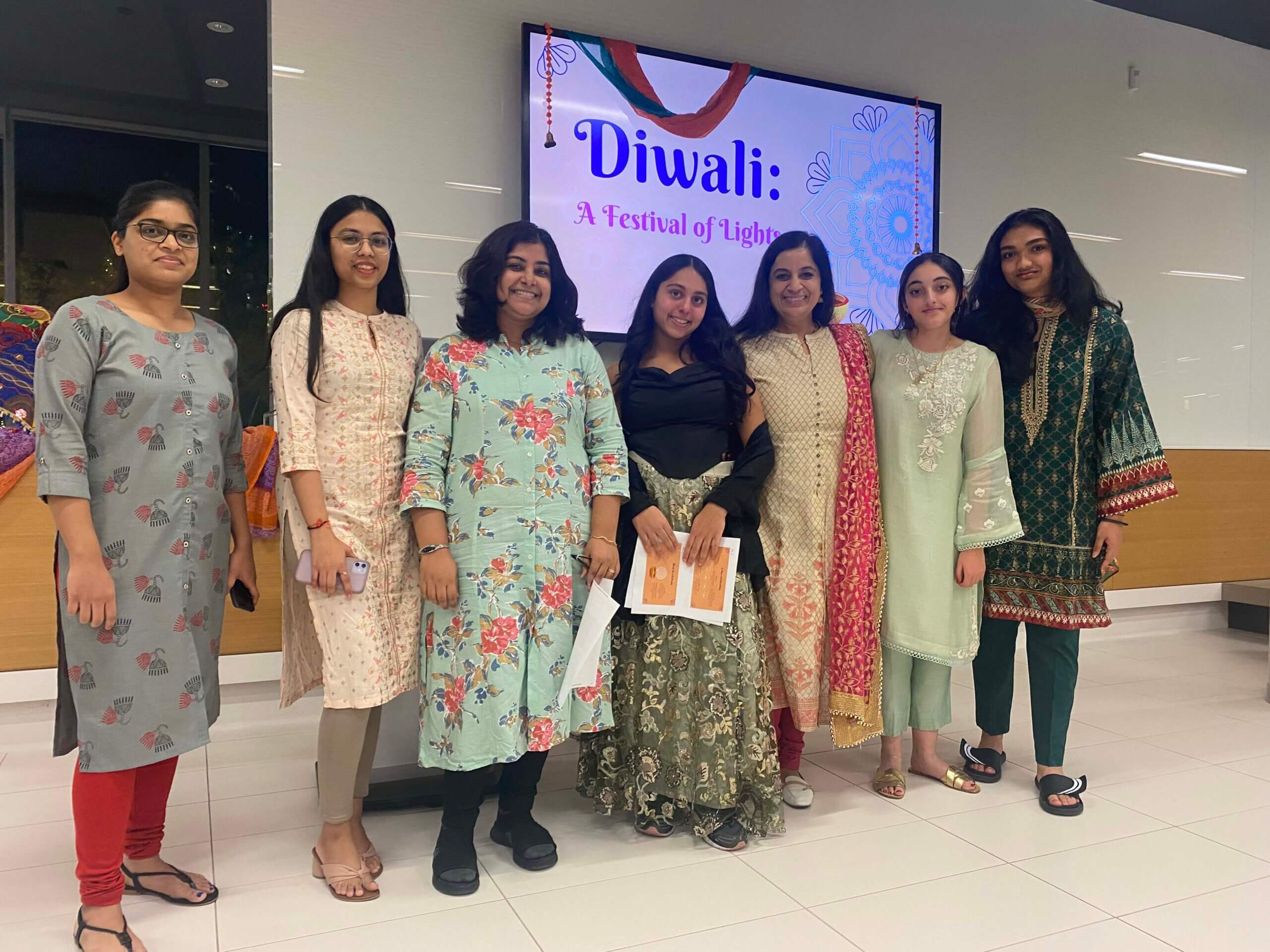 Seven women stand together indoors, dressed in colorful traditional outfits, smiling at the camera. Behind them, a screen displays “Diwali: A Festival of Light.” The setting appears festive and celebratory.