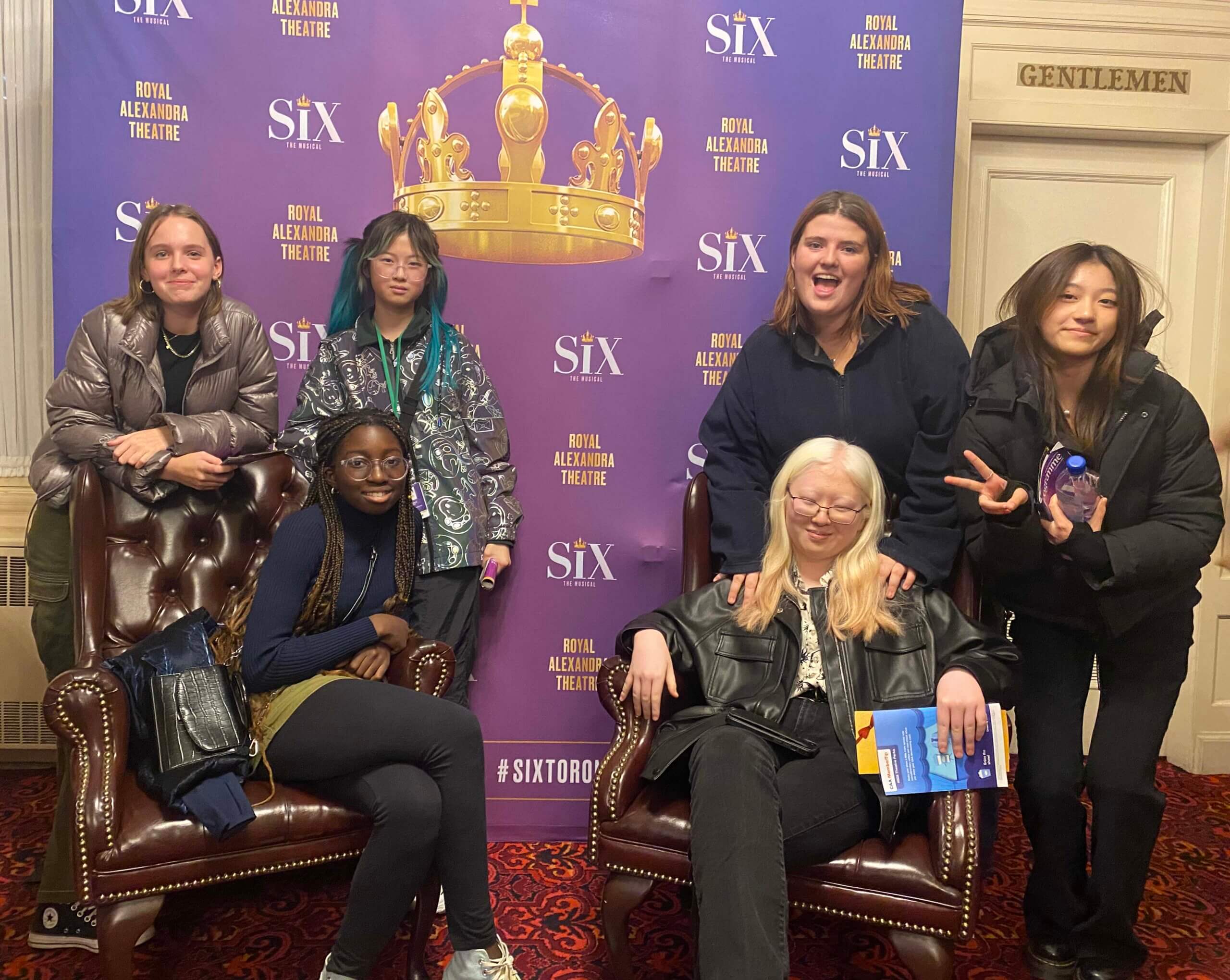 Six young people pose in front of a purple “SIX” musical backdrop at the Royal Alexandra Theatre. Two are seated on brown chairs, while four stand behind them, all smiling or making peace signs.