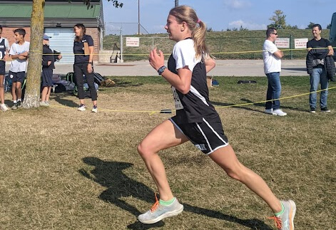 A female runner in athletic gear is sprinting outdoors on grass, while several people stand and watch in the background near a building and a tree.