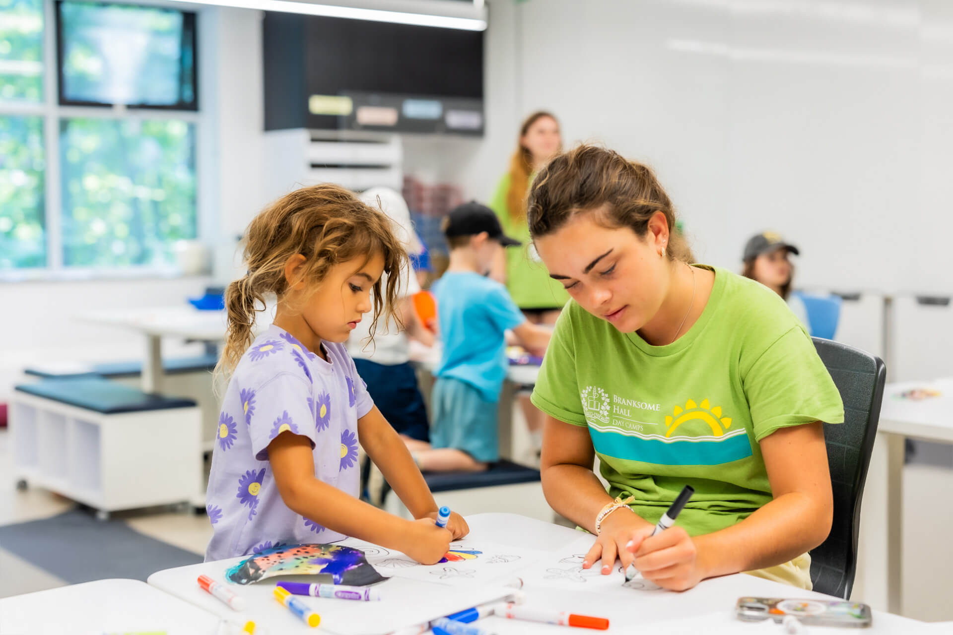 A young woman and a little girl draw together at a table in a bright classroom. The girl uses markers while the woman colors with a pen. Other children and an adult are visible in the background.
