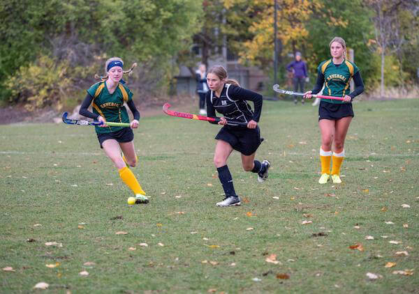 Three female field hockey players compete on a grassy field. One player in black runs with the ball, pursued by two players in green and yellow uniforms. Autumn trees are visible in the background.
