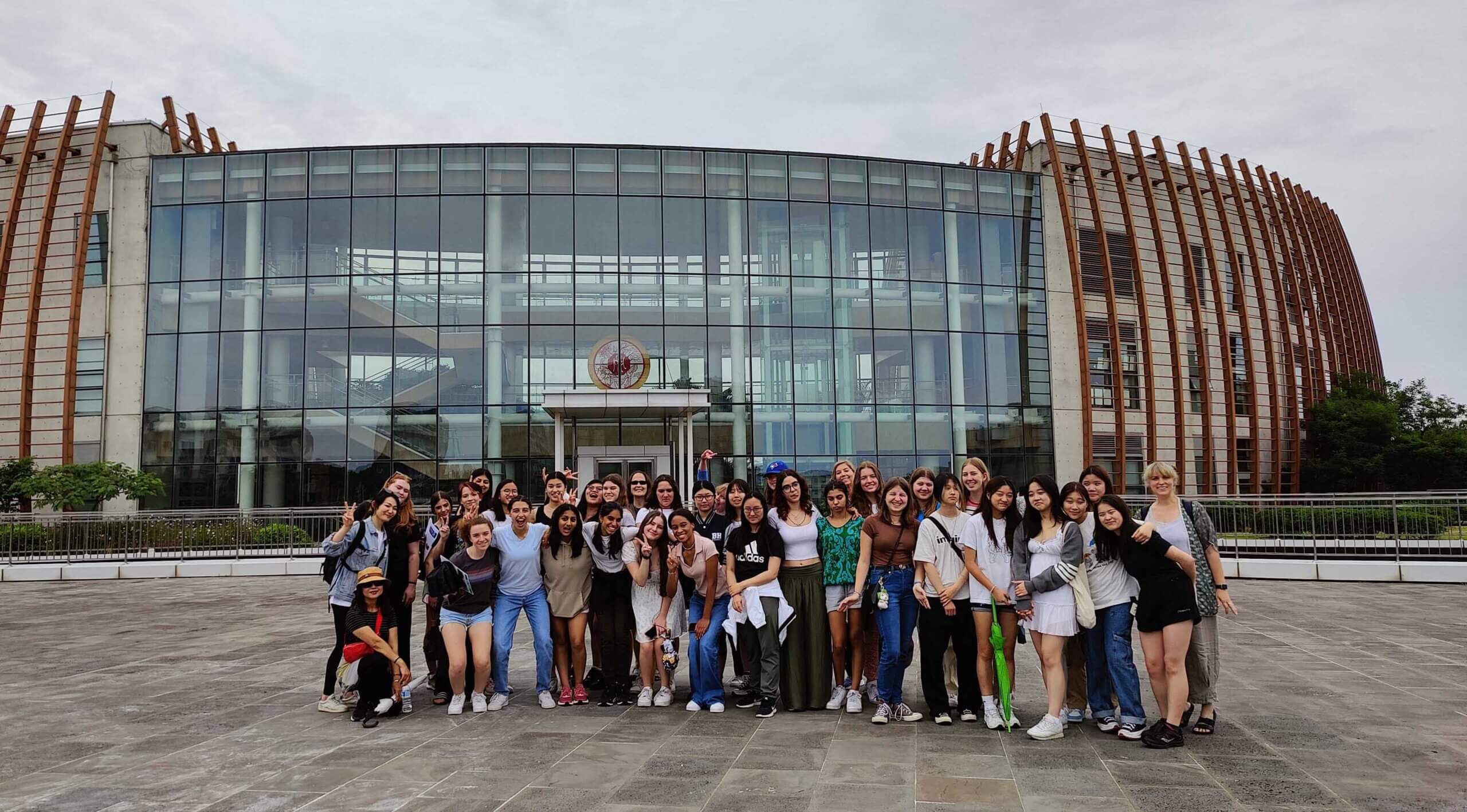 A large group of smiling people pose together in front of a modern glass building with a circular emblem above the entrance and distinctive brown vertical beams on the sides.