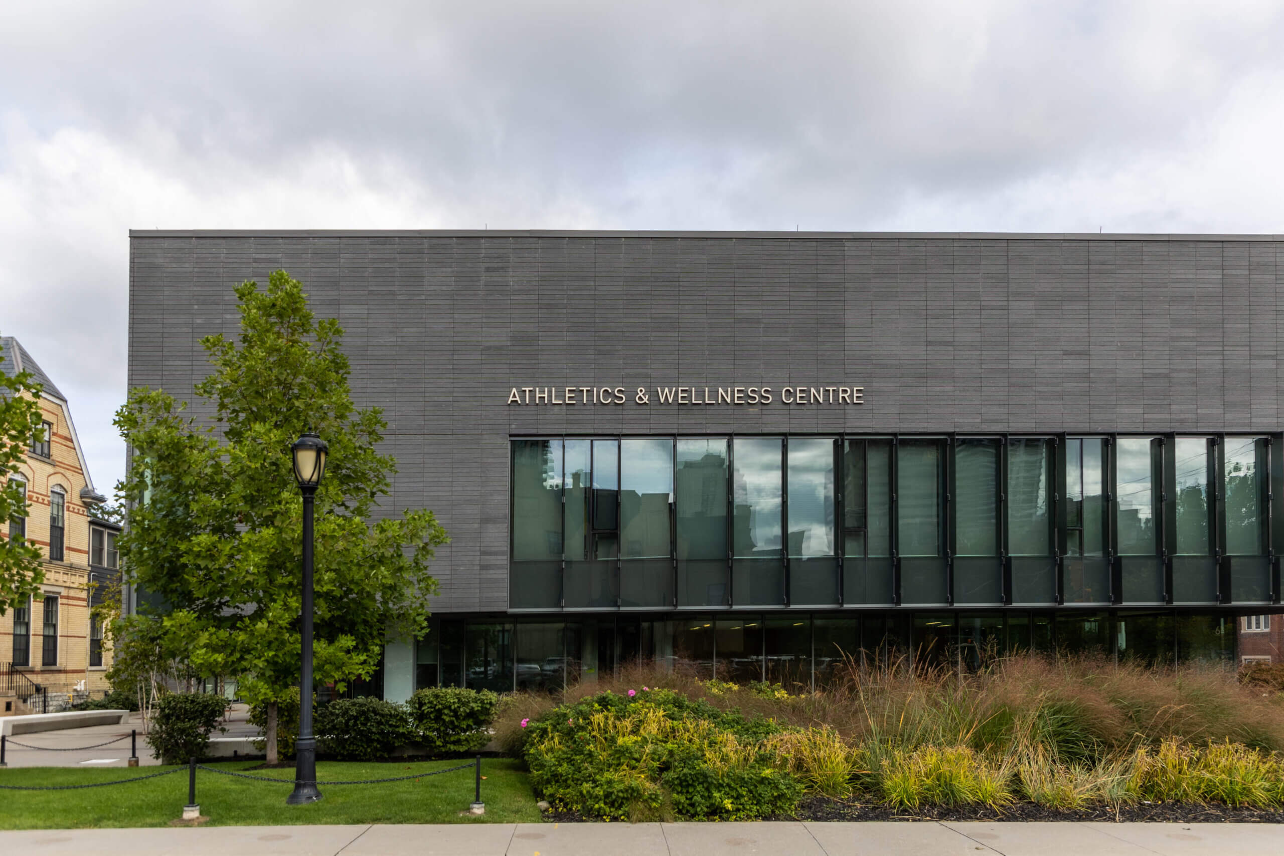 A modern building with large windows and a sign reading "ATHLETICS & WELLNESS CENTRE" is surrounded by trees, shrubs, and a paved walkway under a cloudy sky.