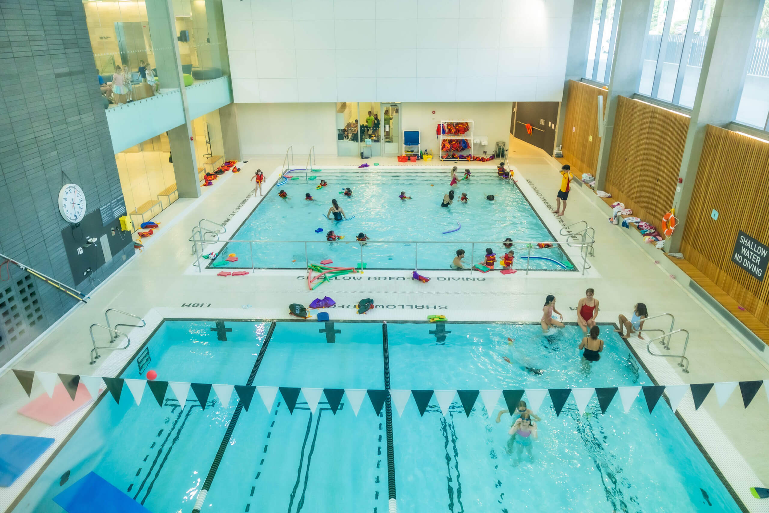 Indoor swimming pool complex with multiple pools; children and adults swim and play, some taking lessons. The area is bright, with large windows, pool equipment, and a clock on the wall. People are supervised by instructors.