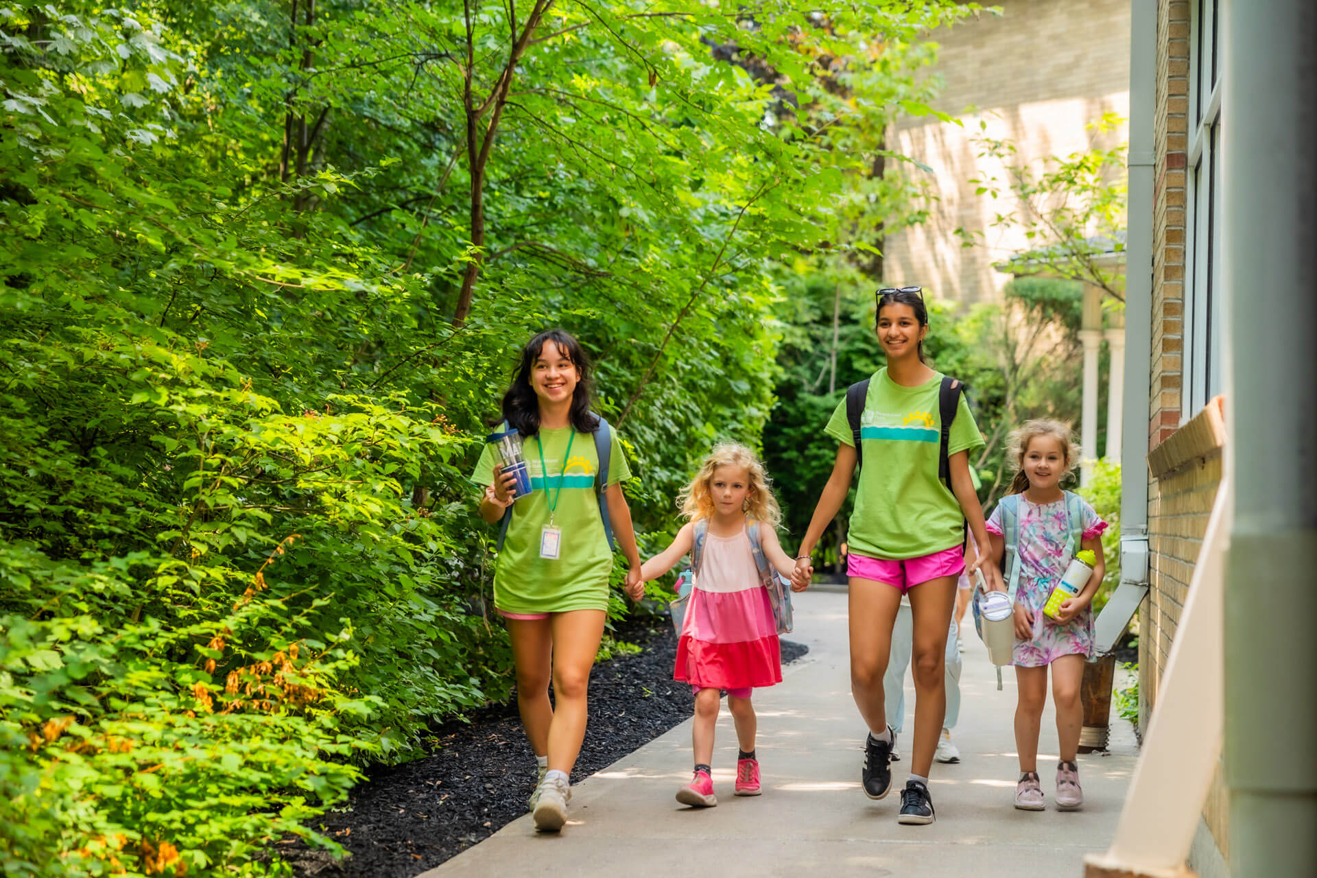Two adults and two young children walk together on a shaded path surrounded by green foliage. The adults wear green shirts and shorts, while the children wear colorful summer clothes. All appear cheerful and engaged.