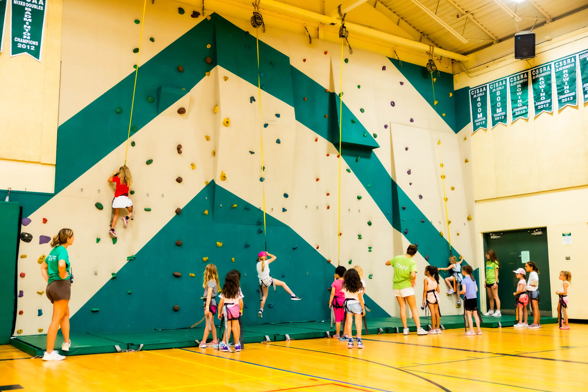 Children and adults in a gym watch as several kids climb an indoor rock climbing wall with colorful holds; some children wait their turn, while staff members supervise.