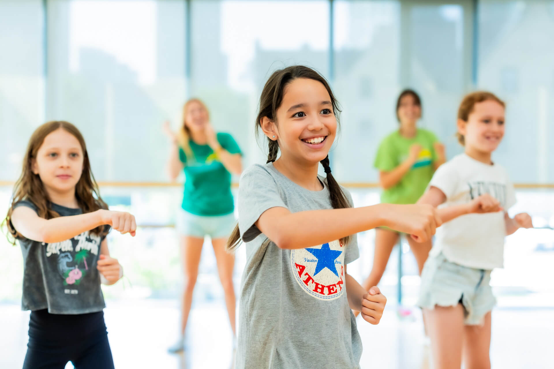 A group of children smile and perform dance moves in a brightly lit room with large windows. The focus is on a girl in a gray T-shirt, with other kids and an adult instructor in the background.